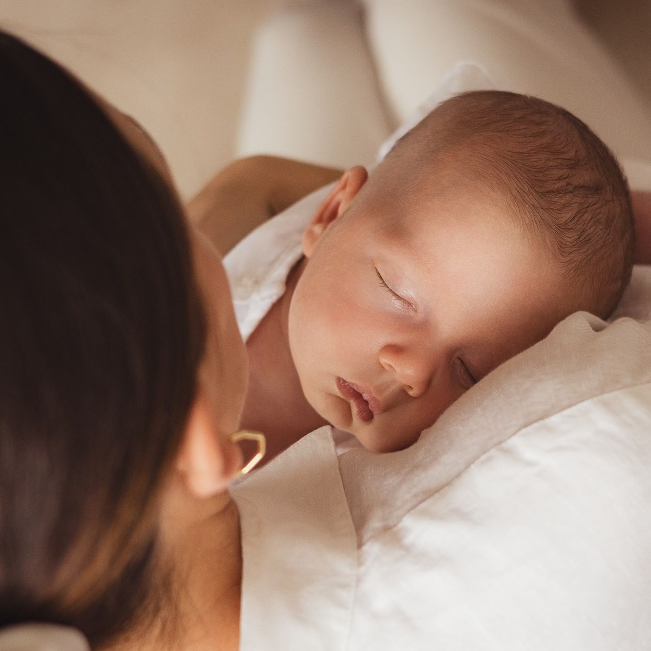 A newborn baby sleeping peacefully in mother's arms, cradled against white fabric in soft warm light.