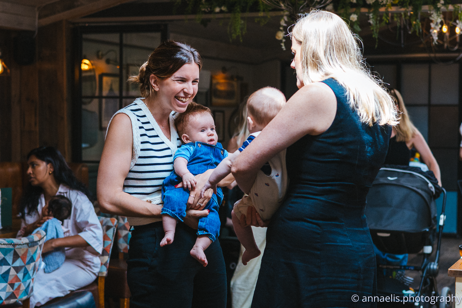 Parents hold their infants while socializing in a dimly lit restaurant space.