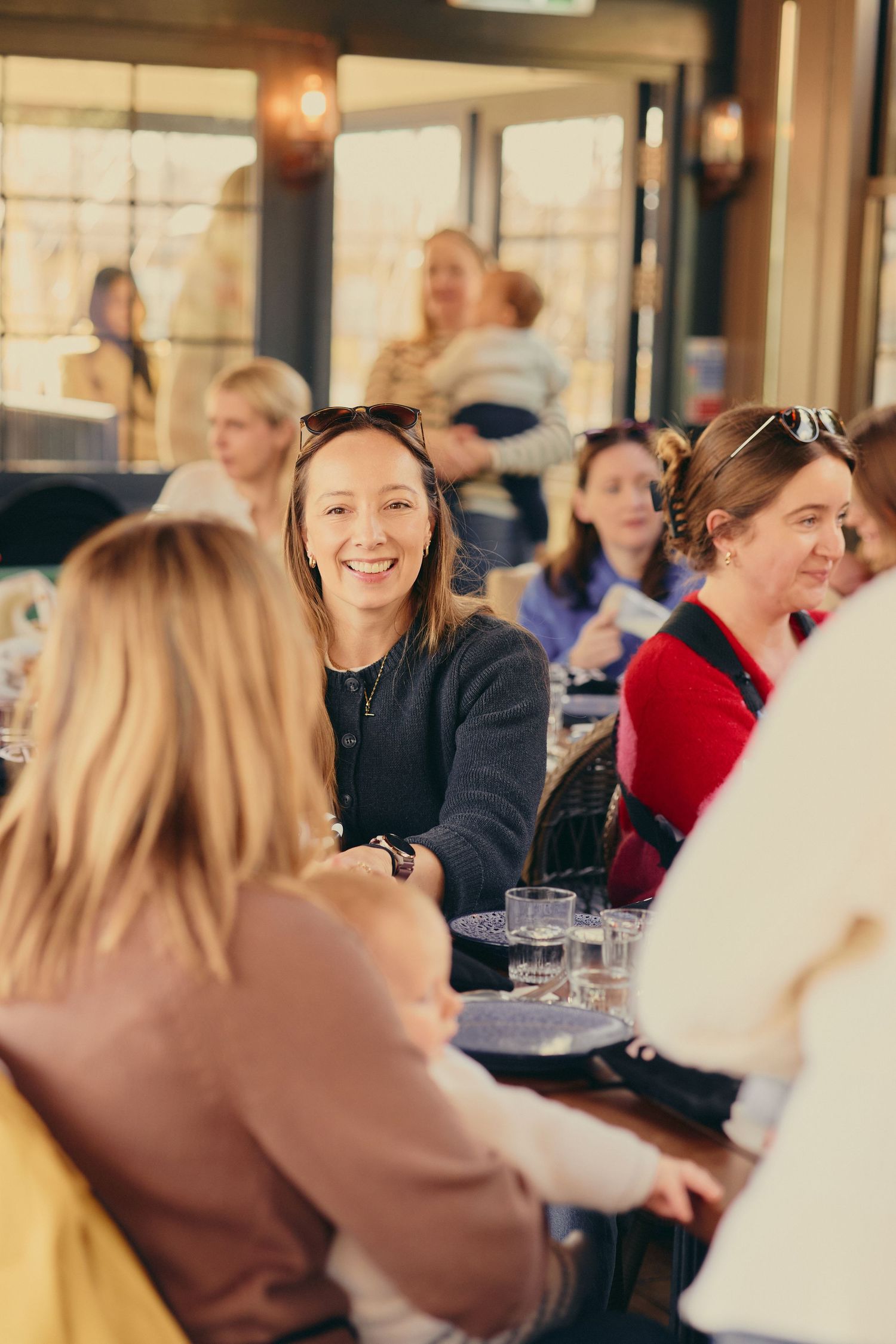 Women smiling and laughing together at a social gathering inside a bright restaurant.