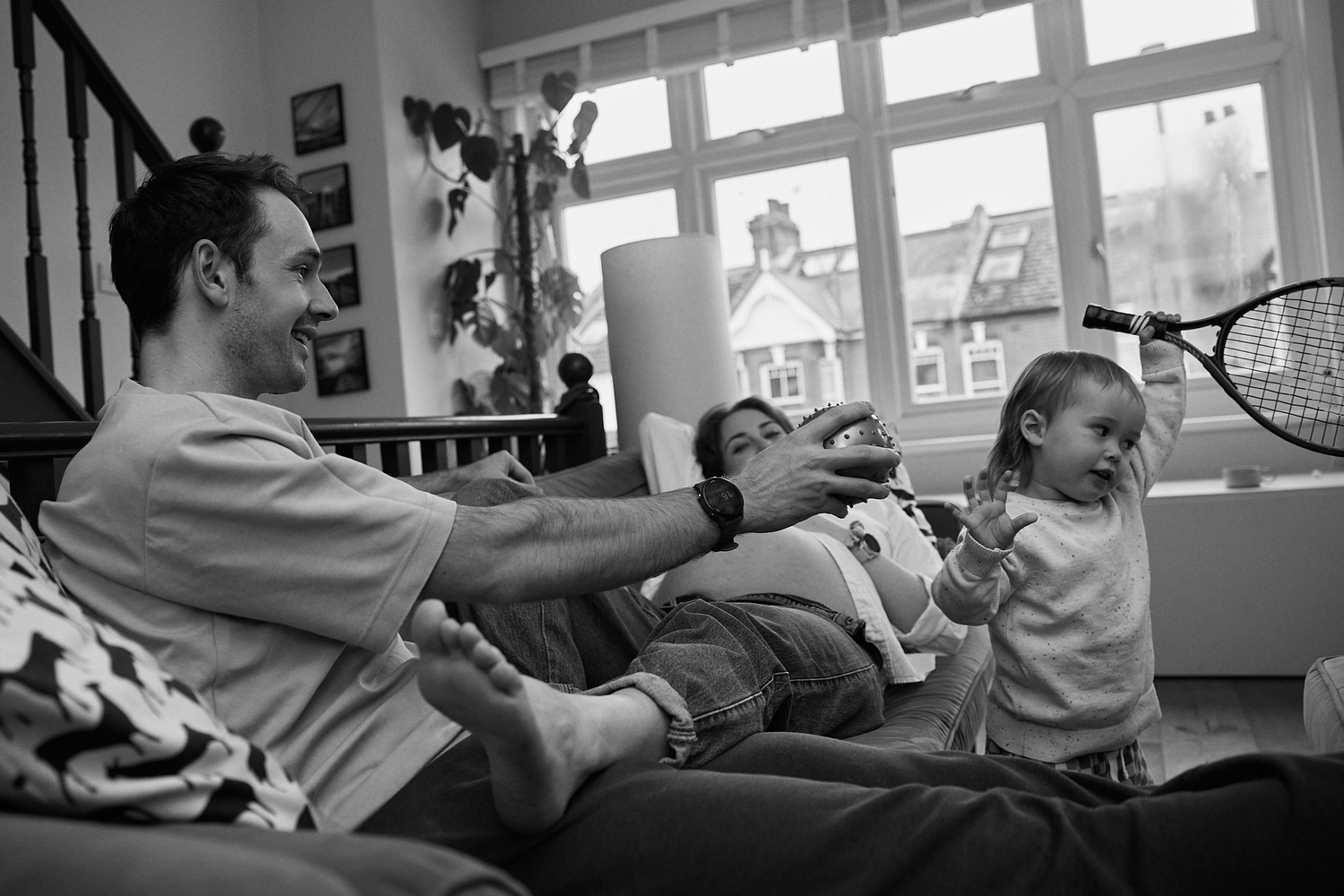 Family relaxing on sofa with man, woman, and toddler playing with tennis racket indoors.