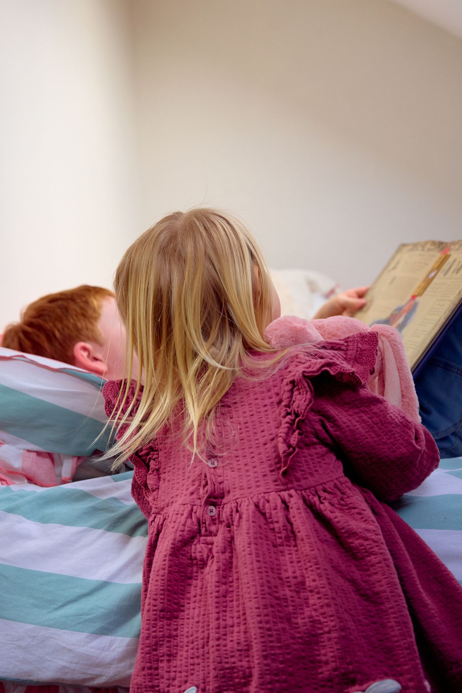 A young girl in a pink dress reads a book with a red-haired child on a striped bed.