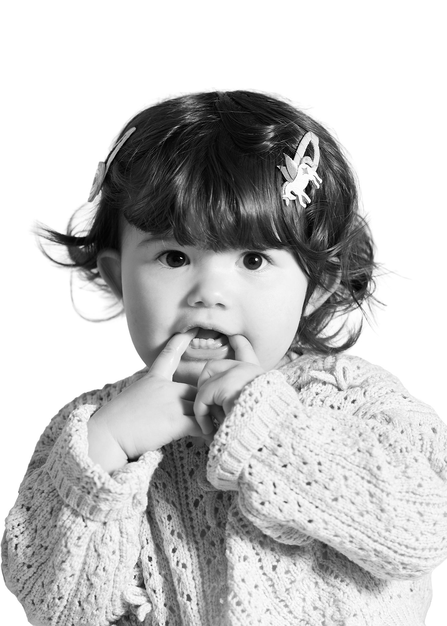 Black and white portrait of a cute toddler girl with hair clip, wearing knit sweater, finger near mouth.