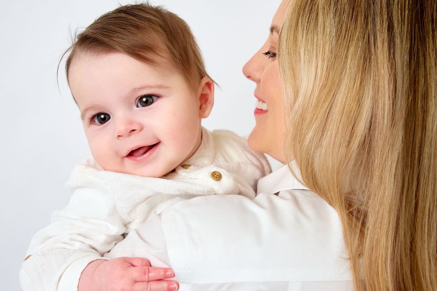 A smiling baby in a white outfit being held by a blonde woman against a white background.
