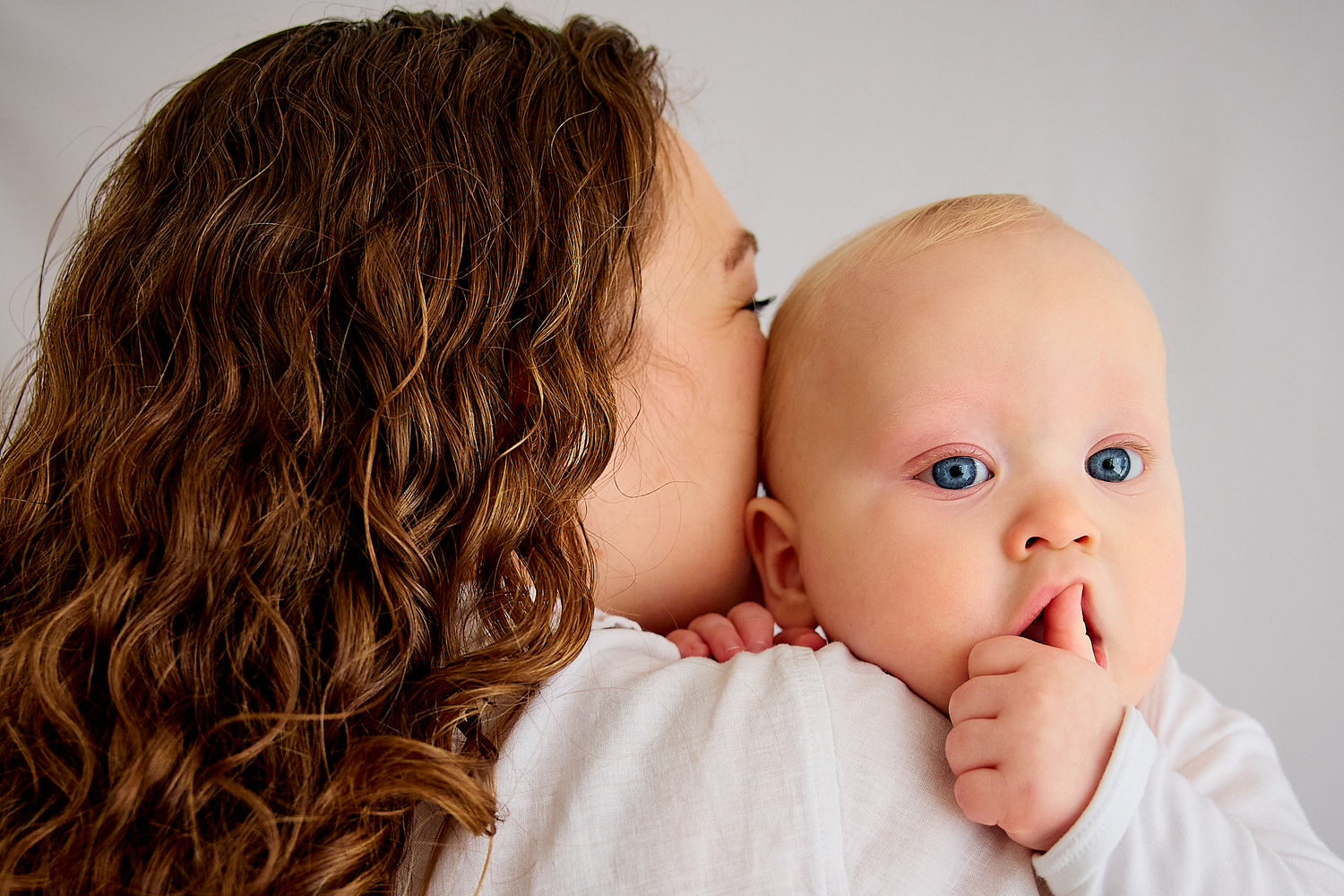 A mother with curly brown hair kissing her blue-eyed baby on the cheek against a white background.
