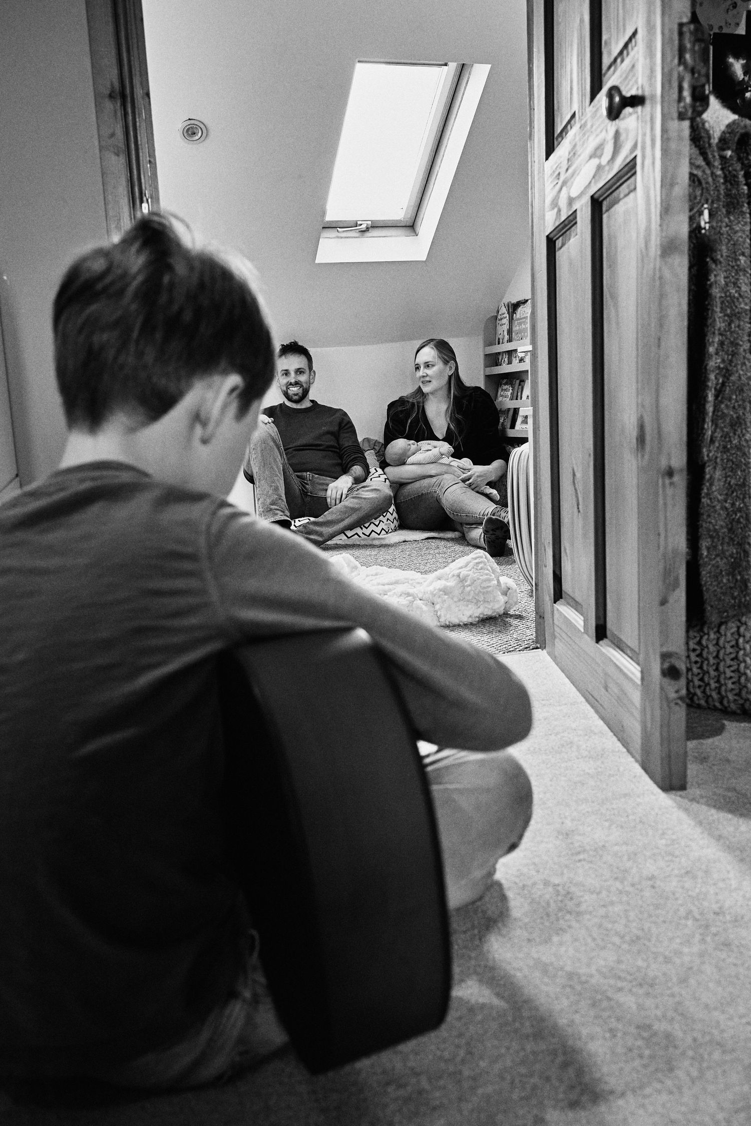 Boy playing guitar for smiling parents holding newborn baby in cozy attic room.