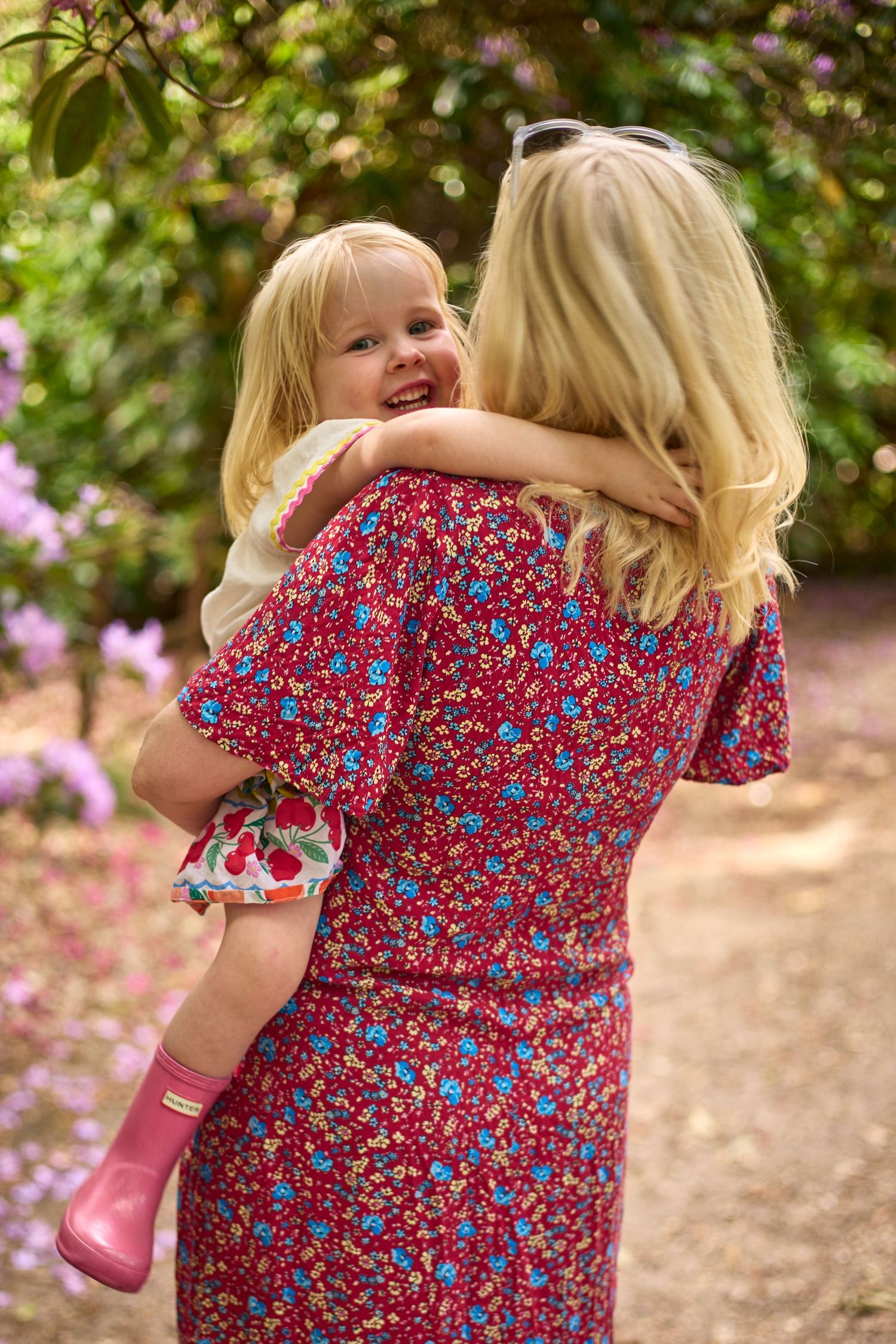 Blonde woman in red floral dress holding smiling toddler girl in pink boots, hugging outdoors in a lush garden.