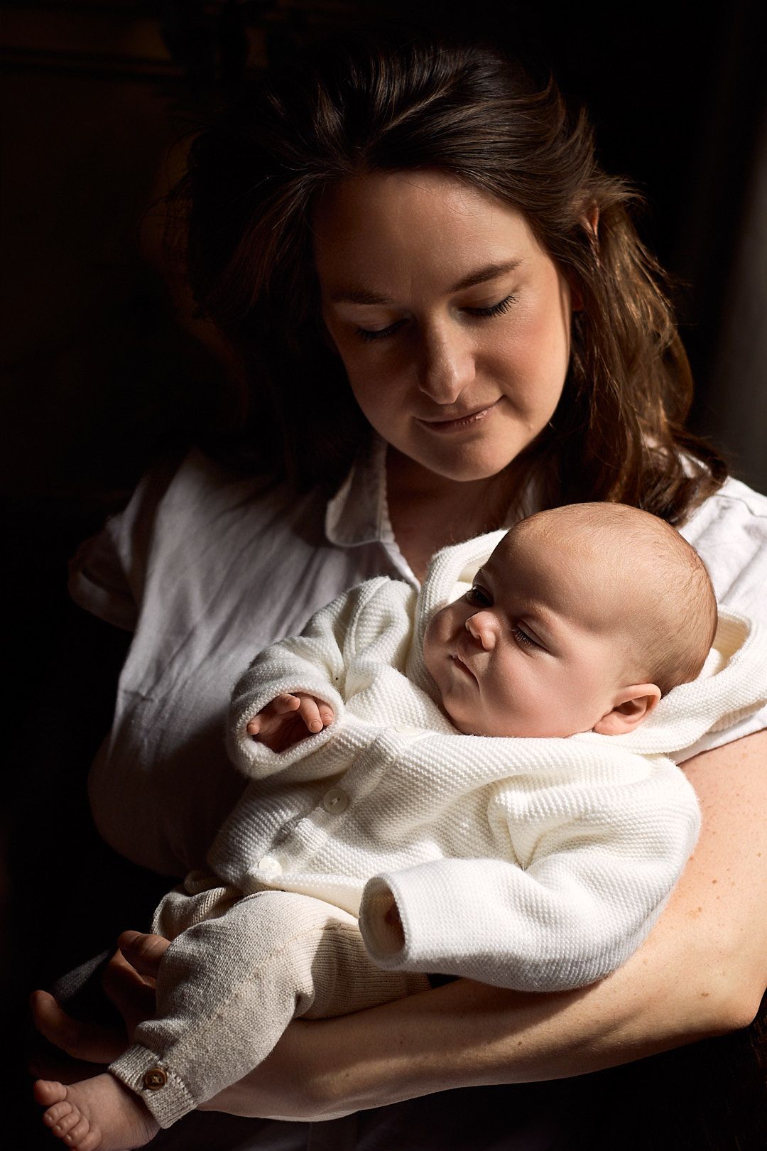 A peaceful portrait in dramatic lighting shows a sleeping infant in white clothing being cradled.