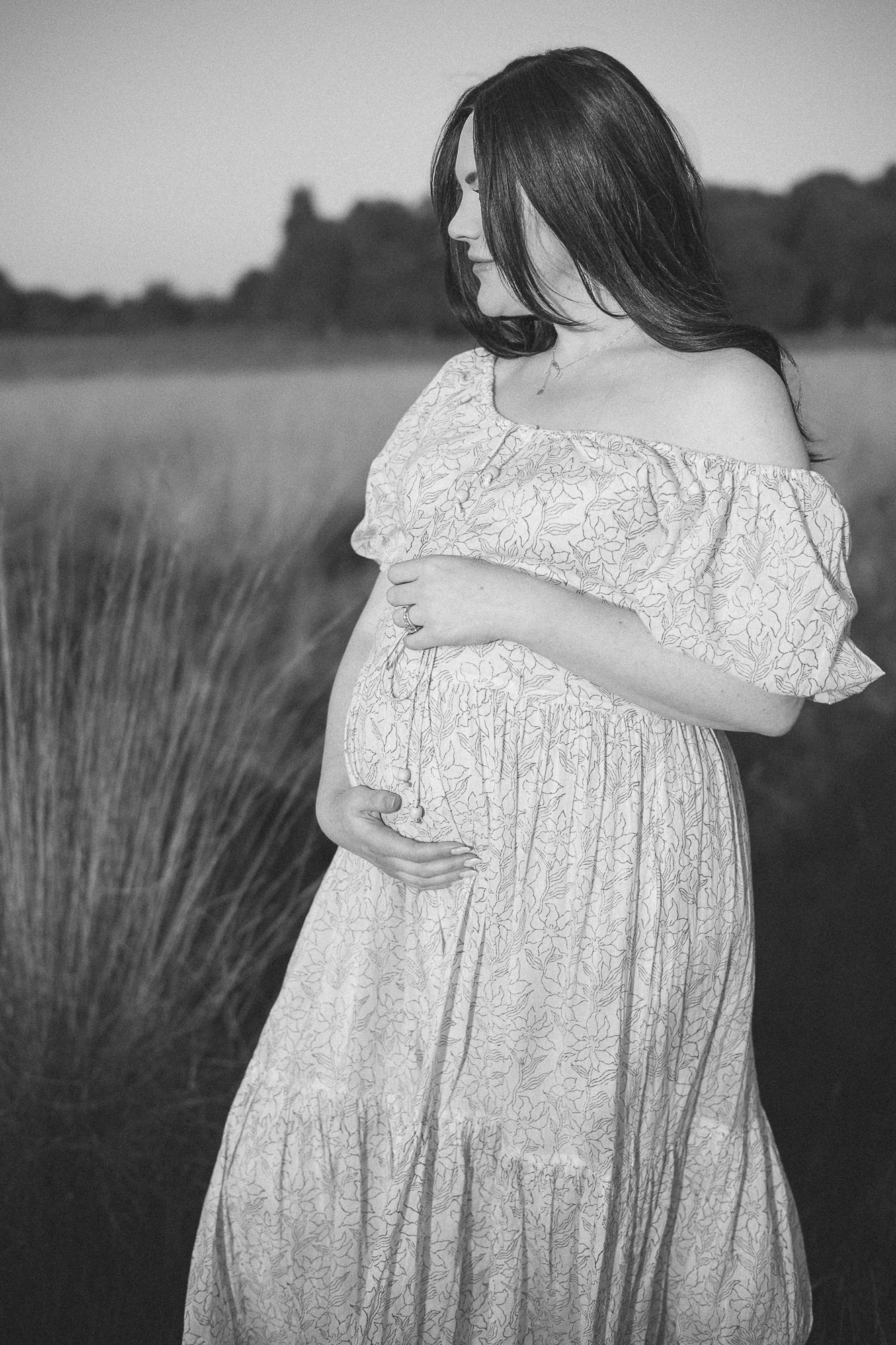 Black and white maternity photo of woman in flowing lace dress standing in field.