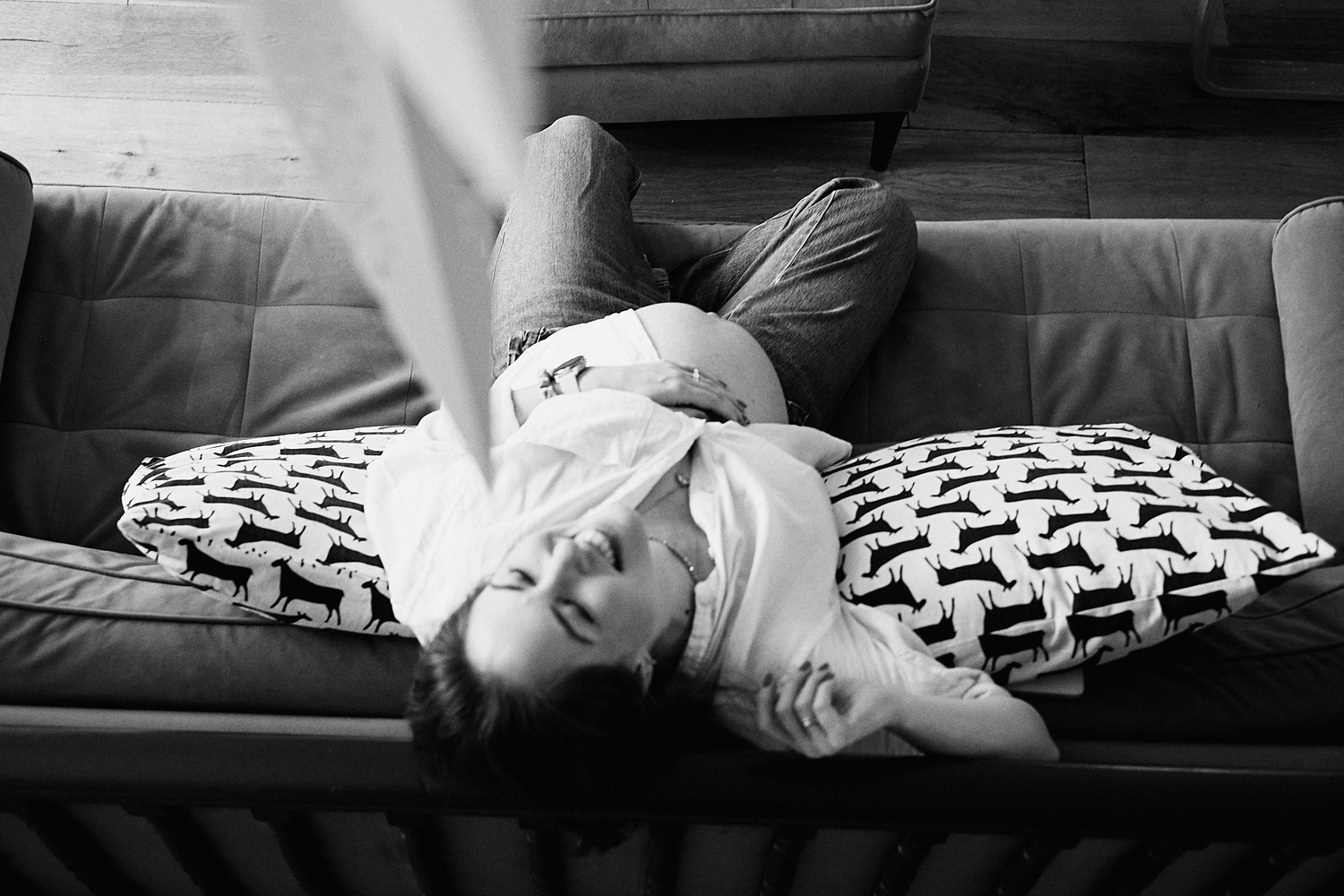 Black and white photo of a woman lying on a leather sofa with patterned pillows, relaxing indoors.