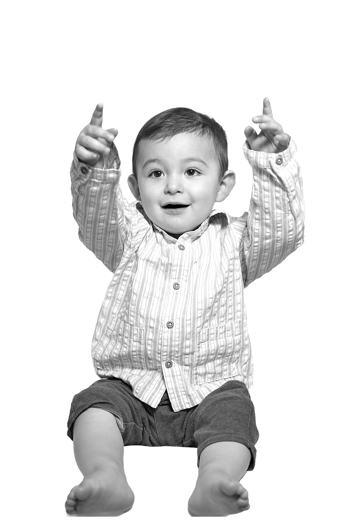 Black and white photo of a happy toddler boy sitting with arms raised and fingers pointing up.