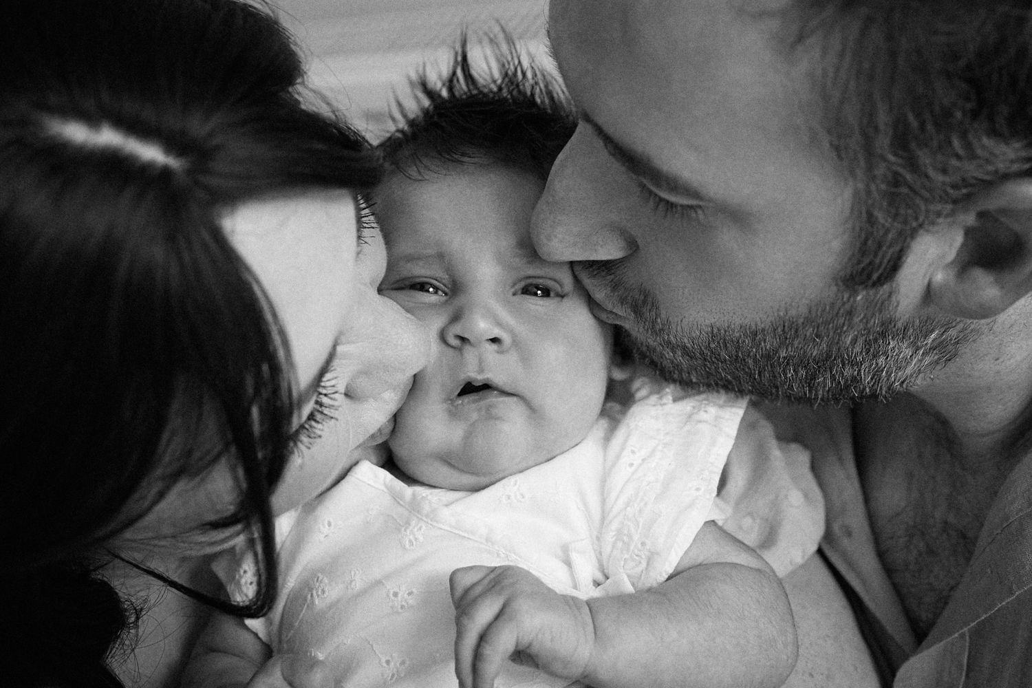 A black and white close-up of parents giving their baby gentle kisses.