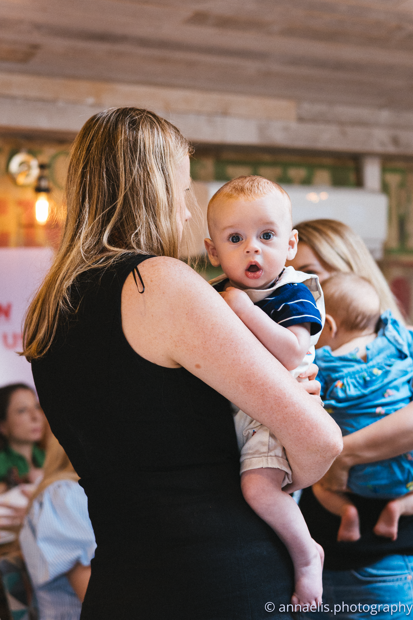 Parents hold their babies during a social gathering in a rustic cafe environment.