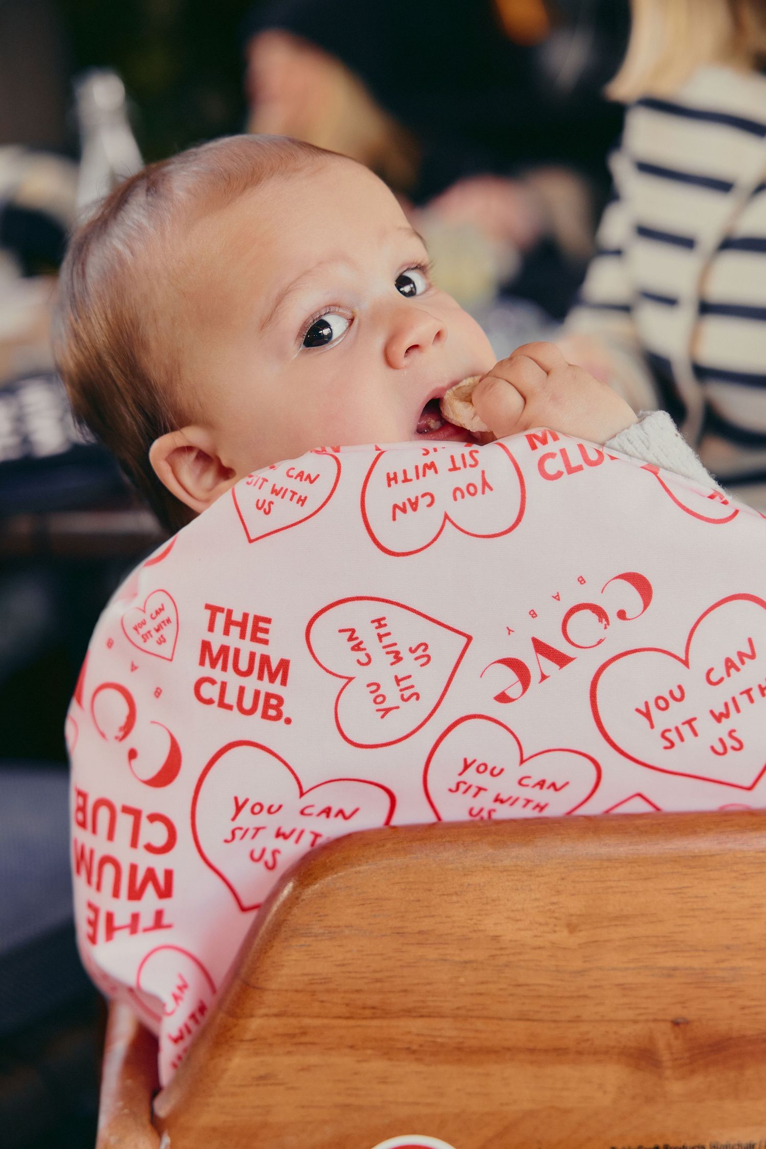 Baby wearing a The Mum Club bib sitting in a wooden high chair chewing fingers.