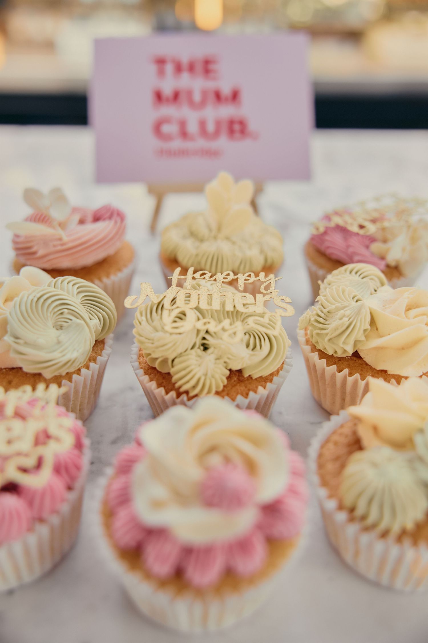 Elegant Mother's Day cupcakes with pink and cream frosting displayed at The Mum Club, featuring floral decorations and festive toppers.