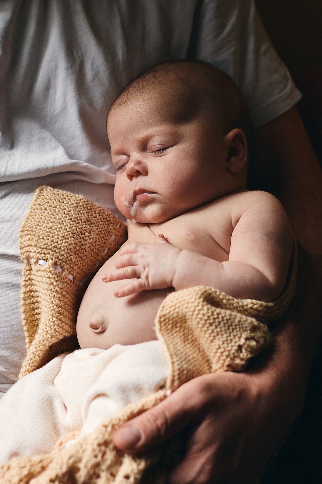 Sleeping newborn wrapped in knitted blanket on white bedding.