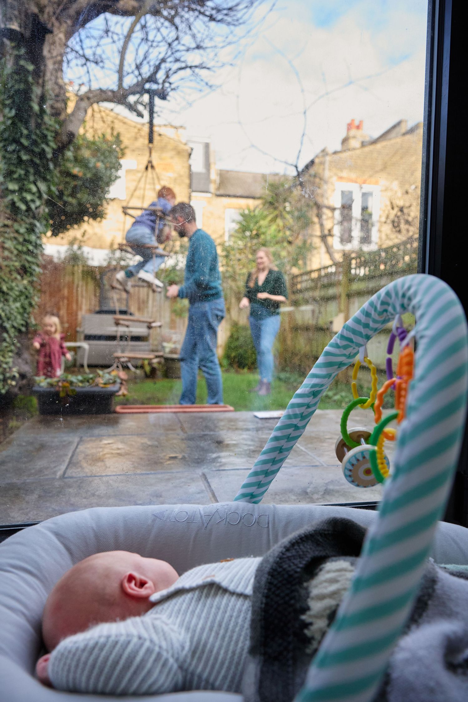 Baby resting indoors while family plays in garden, viewed through glass door on cloudy day.
