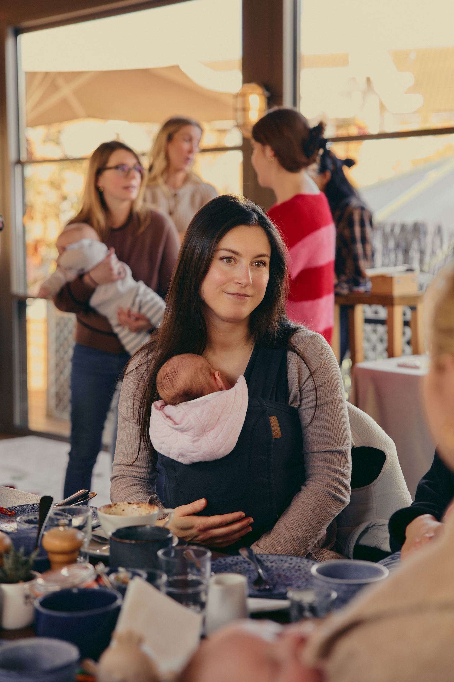 A woman wearing a baby carrier with a newborn sits at a table, with other mothers and babies visible in the background.