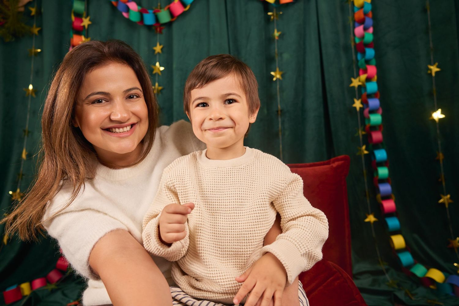 A woman and young boy pose together smiling in front of festive Christmas decorations with colorful garlands and star lights.