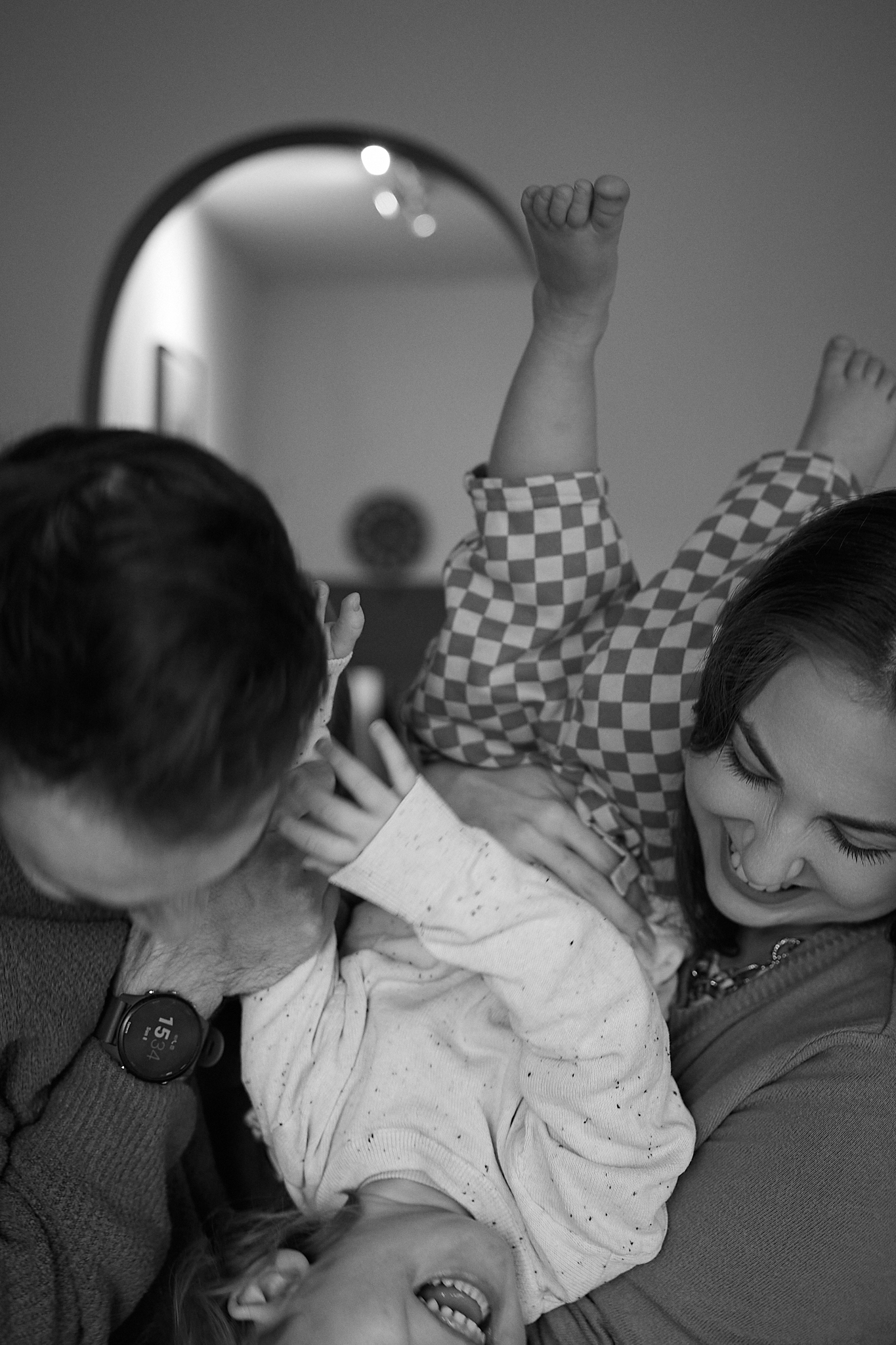 Black and white photo of two parents playfully holding their laughing baby upside down in a cozy home setting.
