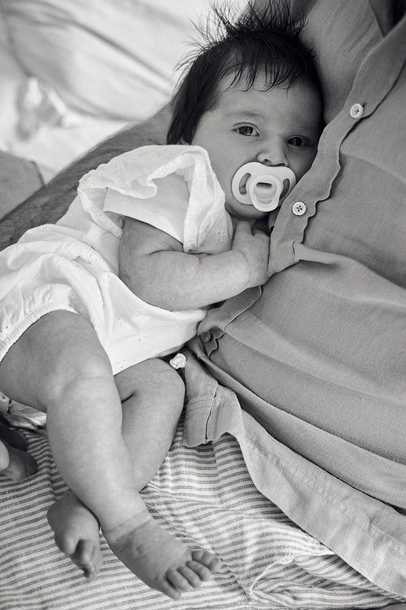 A small baby wearing a white outfit lies on striped bedding with a pacifier.