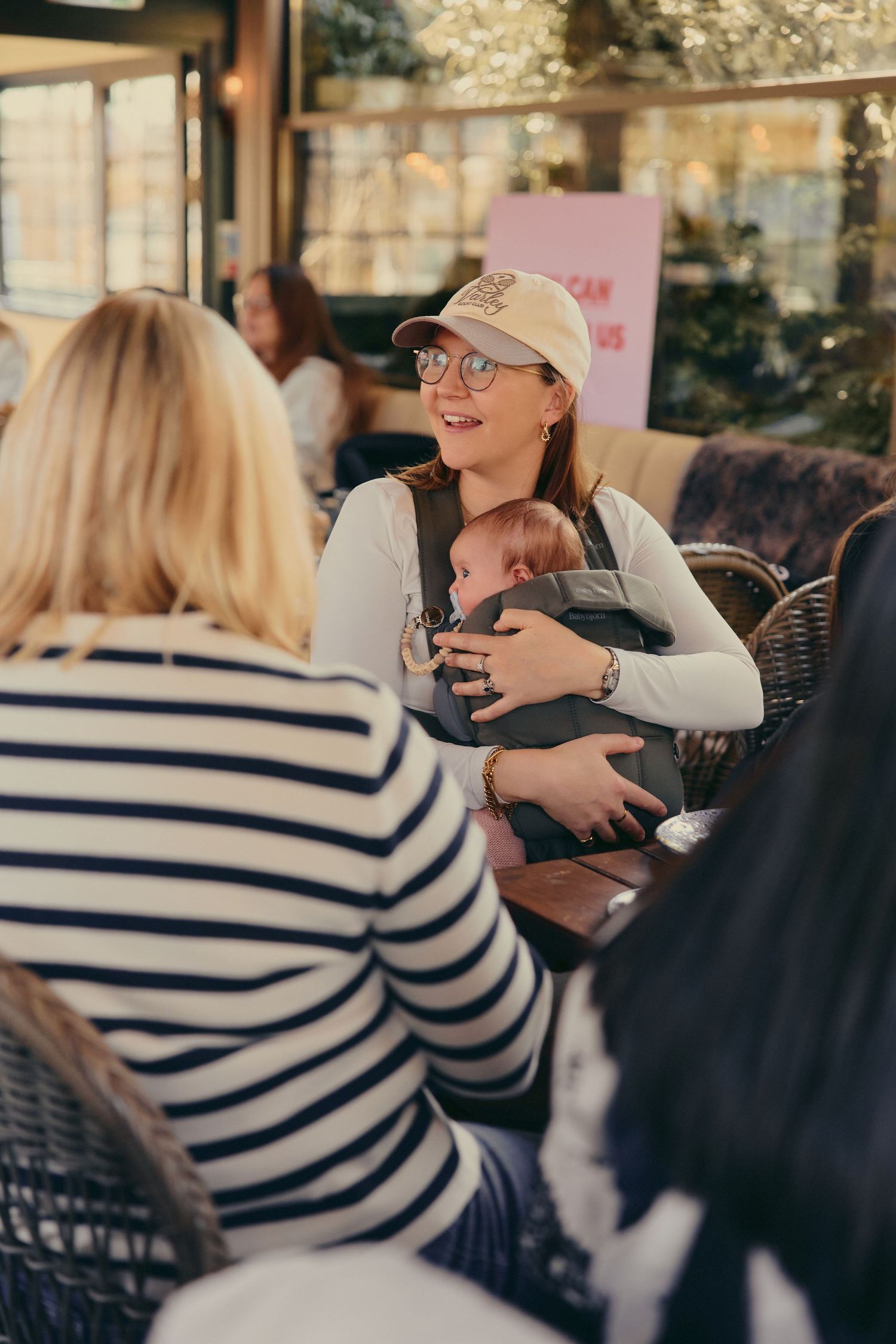 Smiling mother wearing glasses and cap holds sleeping baby in carrier while socializing at a cafe.