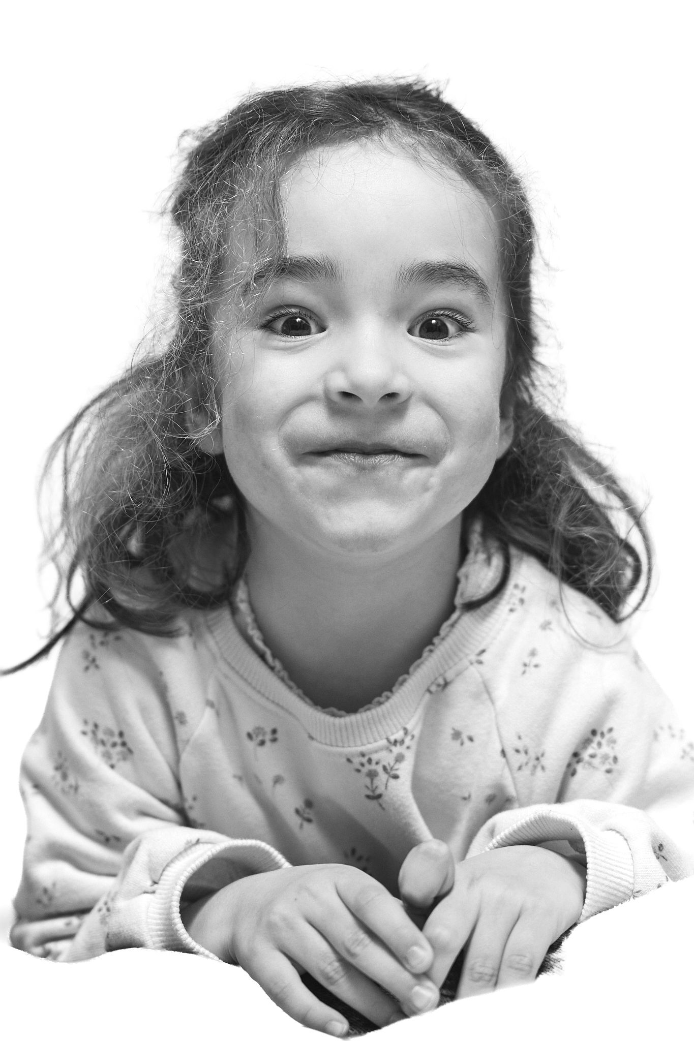 Black and white portrait of a young girl with curly hair smiling playfully at the camera with hands folded.