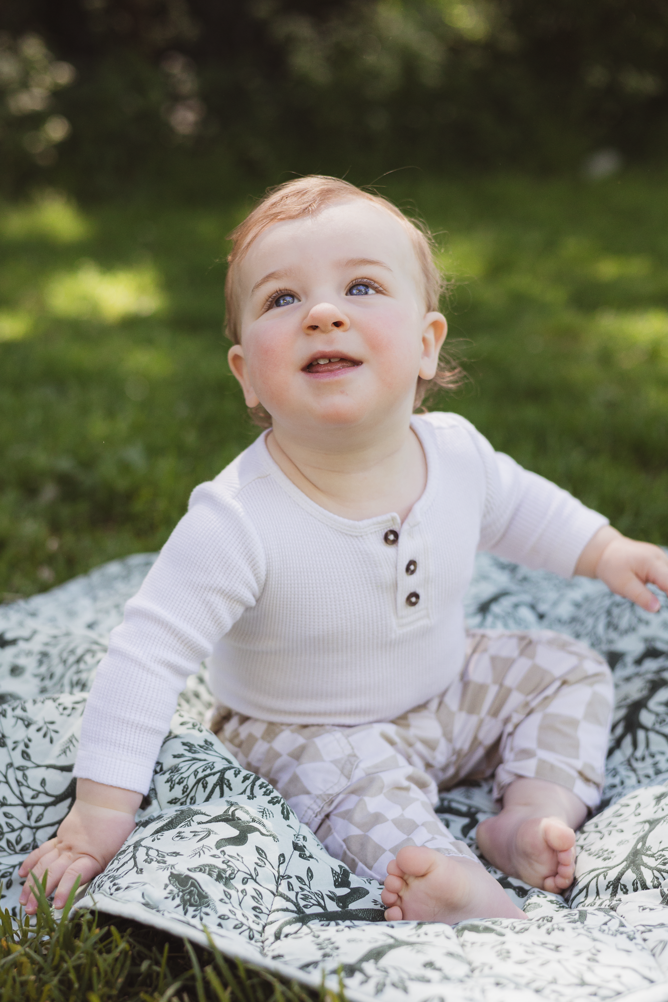 Baby in white henley outfit sits on black and white patterned blanket in garden setting.