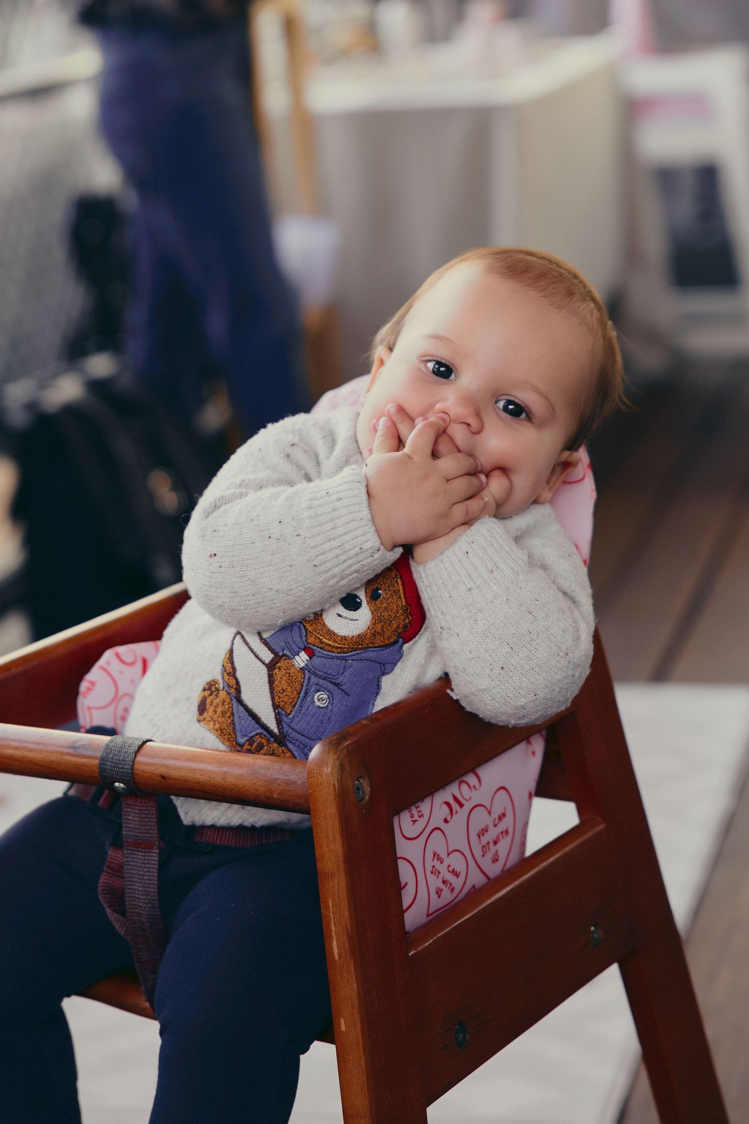 Cute baby sitting in a wooden high chair with hands on face, wearing a white knit sweater with bear design.