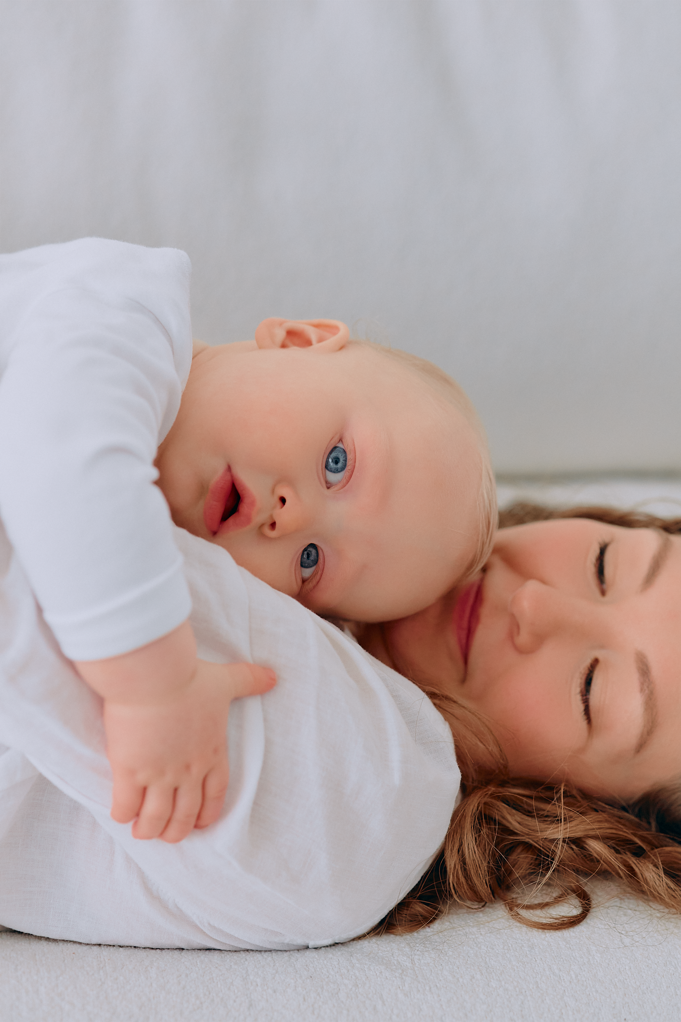 A mother lying down with her blue-eyed baby in white clothing, sharing a tender moment together.