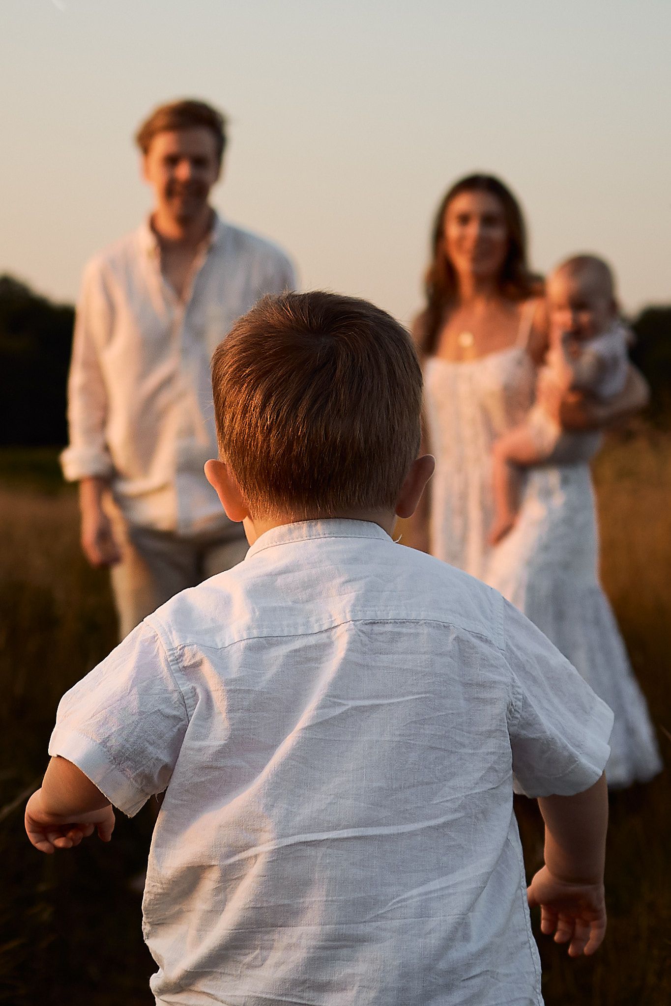 A young child in a white shirt runs towards a family in a sunlit field during golden hour.