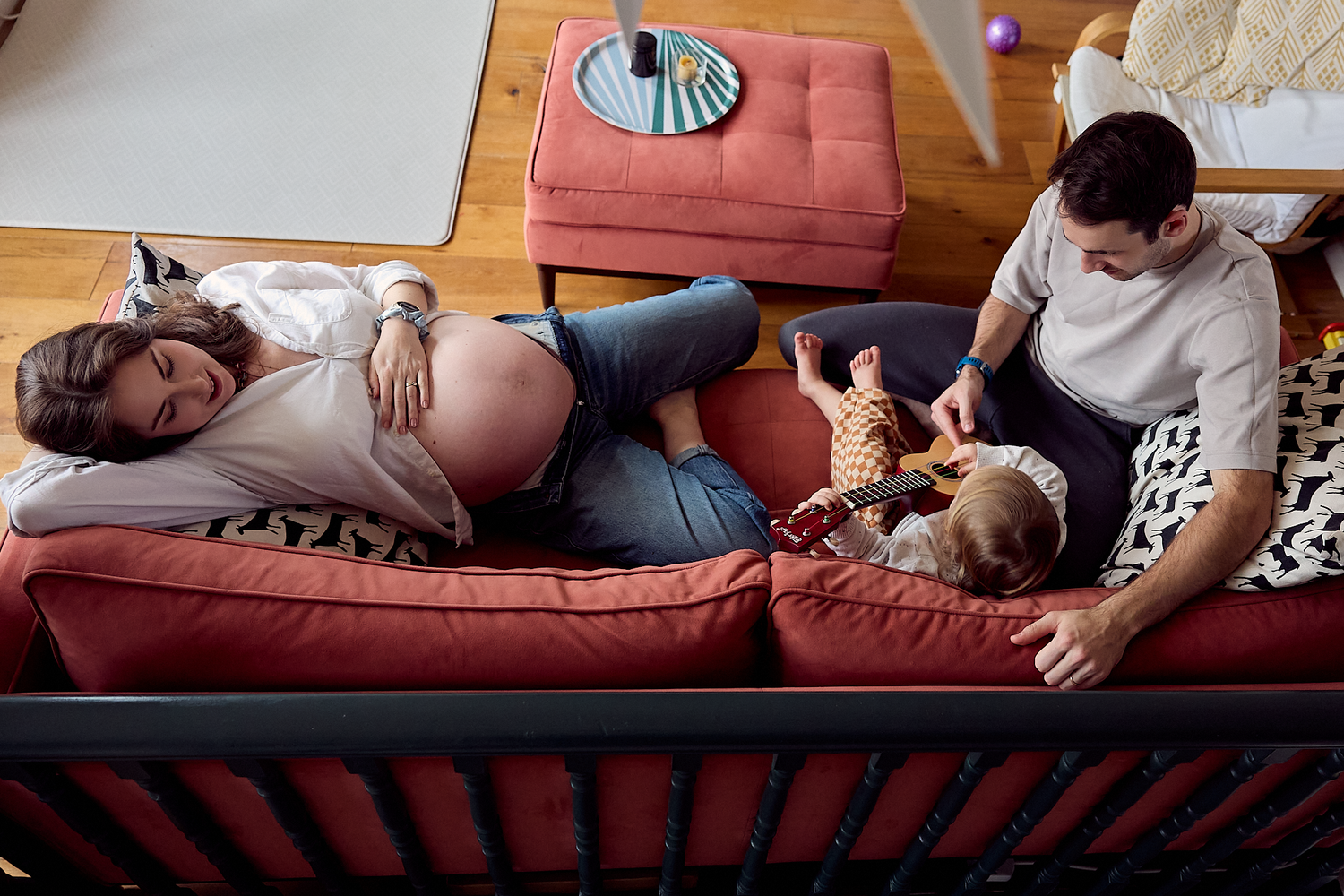 Pregnant woman resting on red sofa with partner and baby viewed from above in living room.