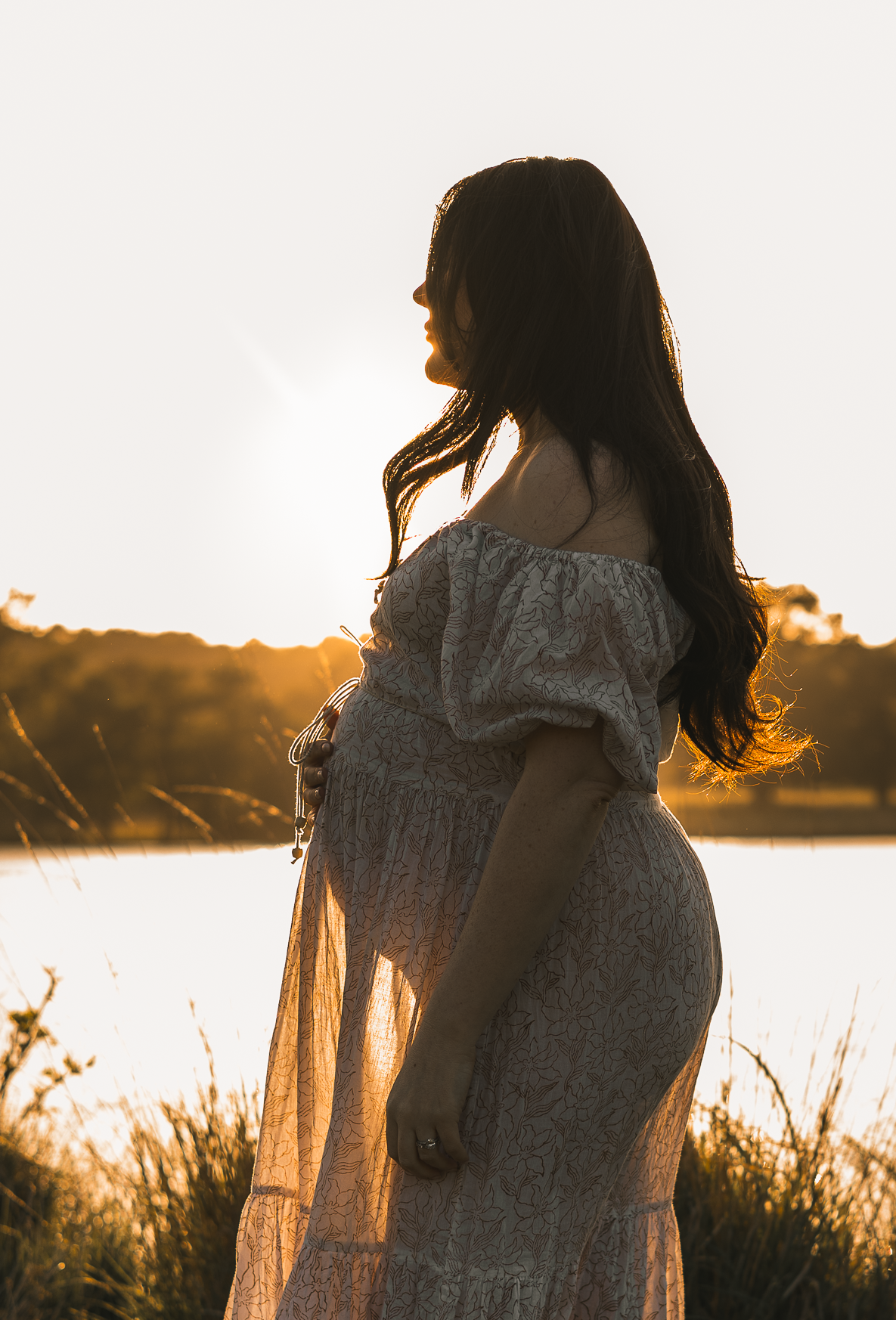 Romantic maternity silhouette in flowing dress captured during golden hour by water.