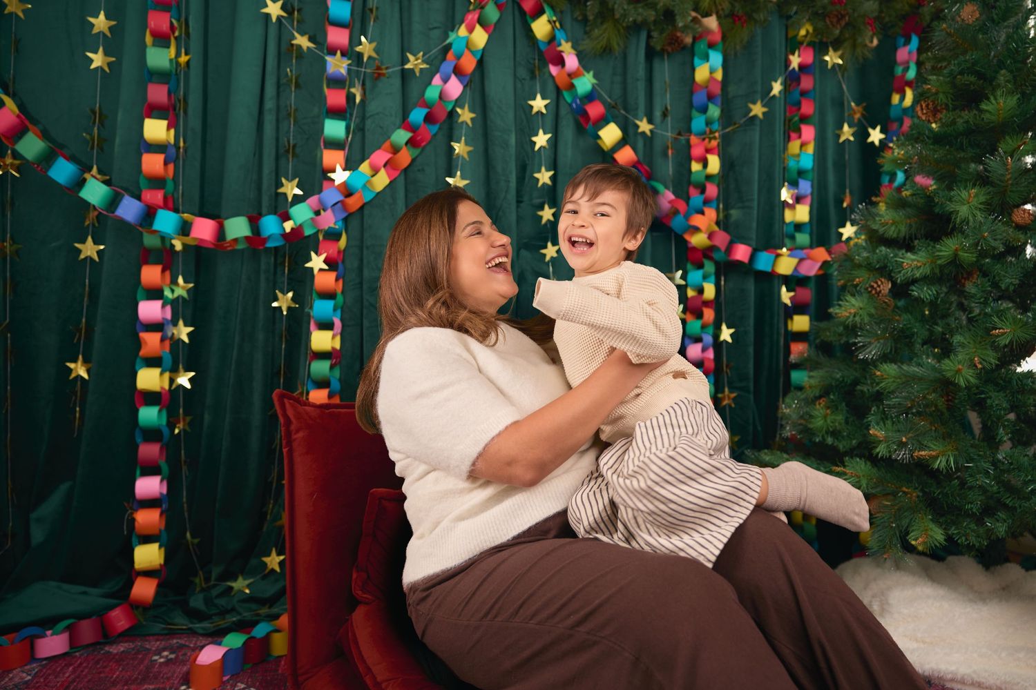 A woman joyfully holds a laughing toddler in front of colorful paper chain garlands and a Christmas tree.