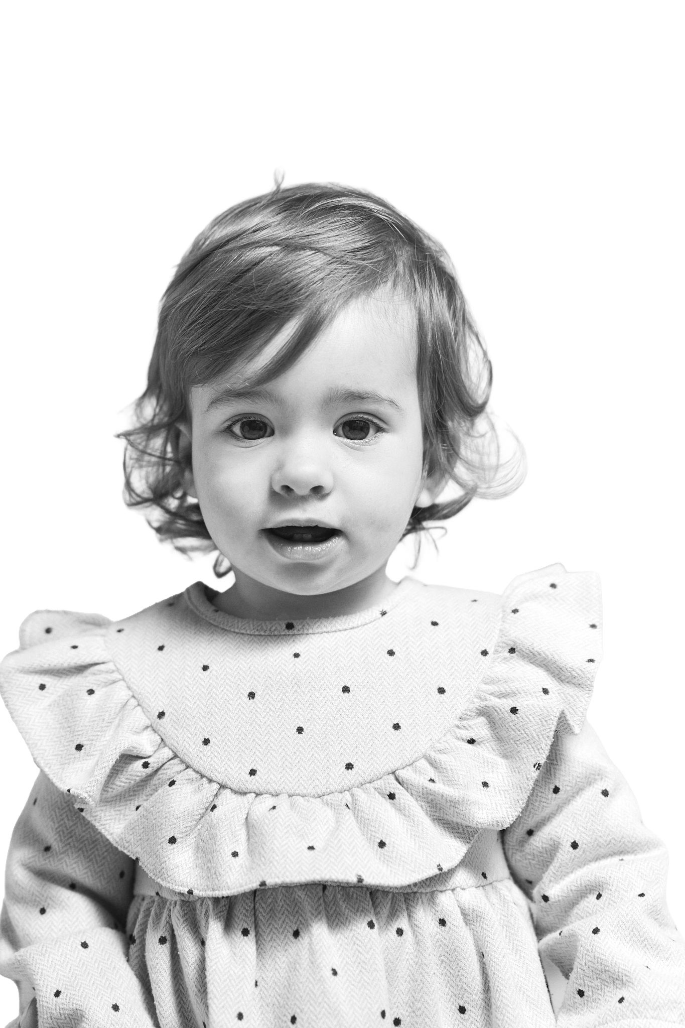 Black and white portrait of a cute toddler girl in a polka dot dress with ruffled collar, looking surprised.