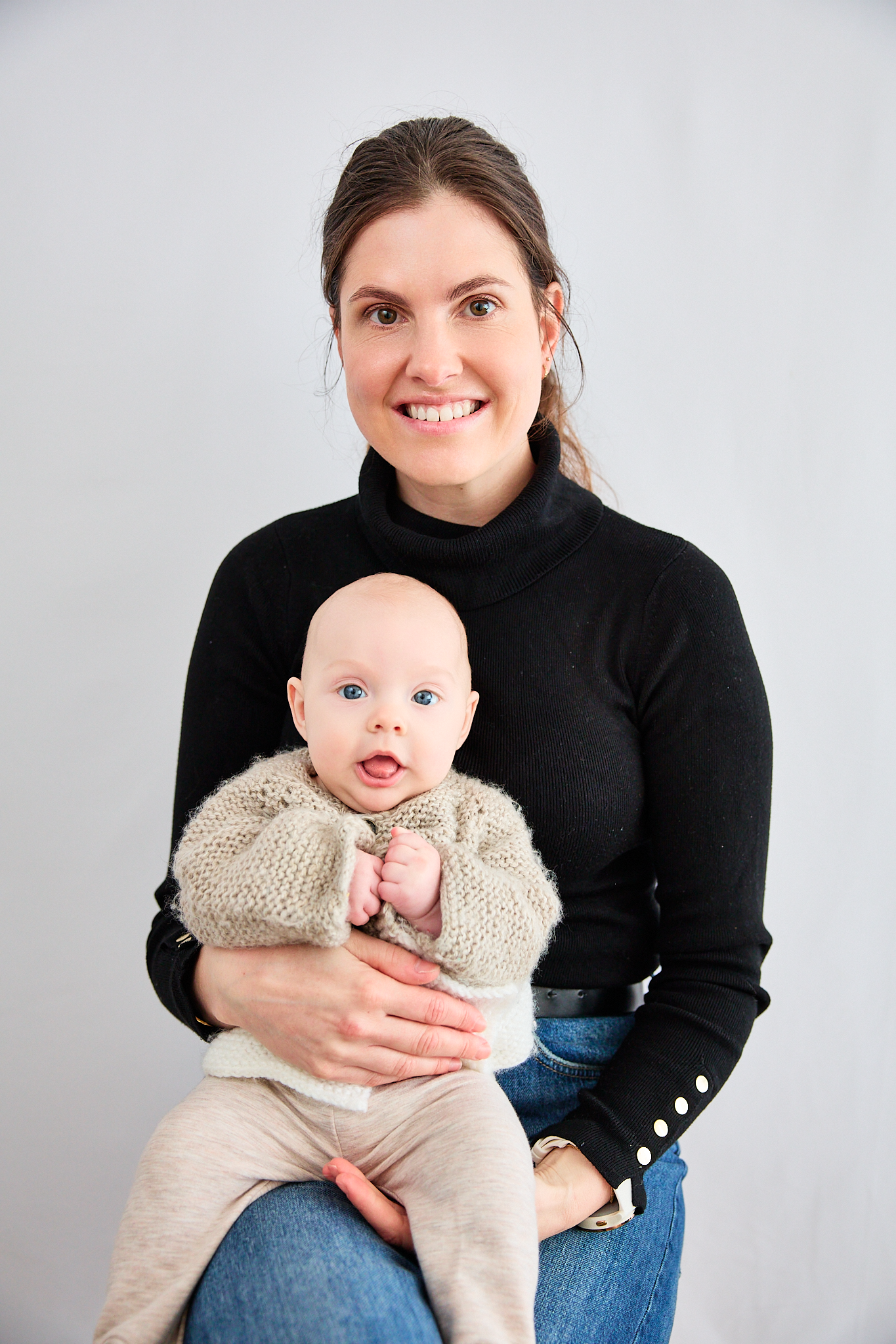 Smiling woman in black turtleneck holding an adorable blue-eyed baby in a knit sweater against a gray background.