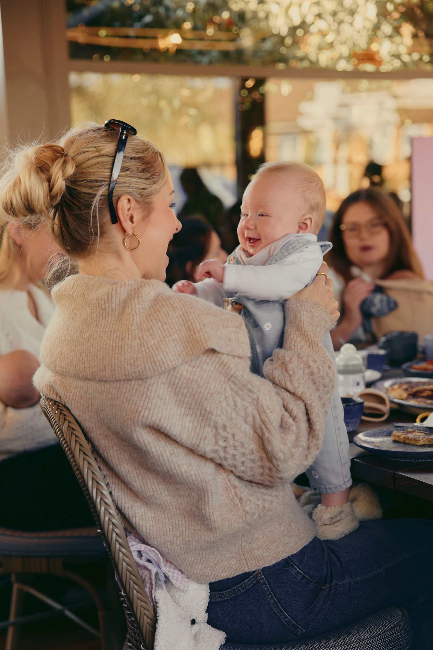 A smiling mother in a beige knit sweater holds her laughing baby at a restaurant gathering.