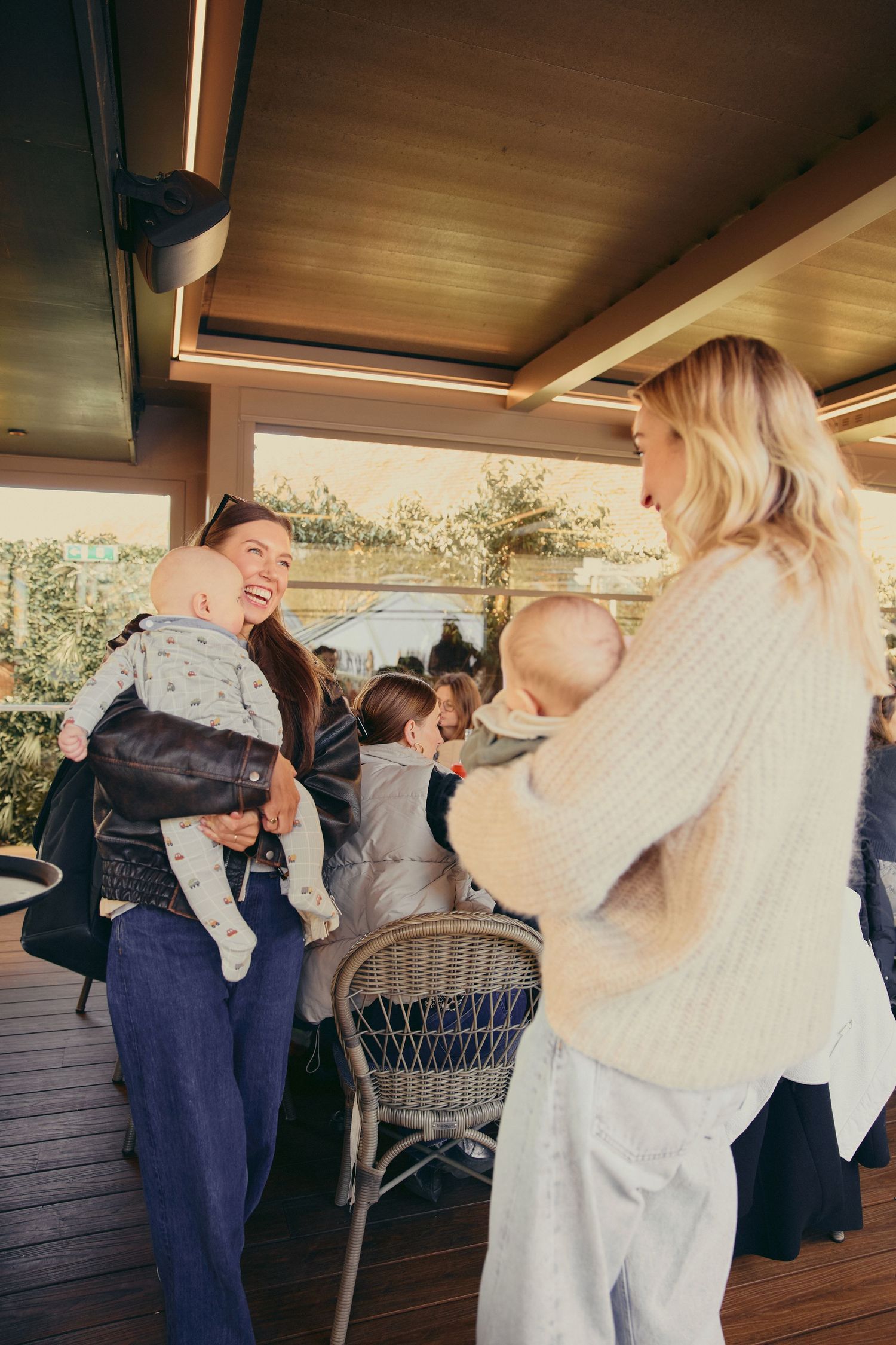 Two smiling mothers holding babies socializing on a sunlit outdoor terrace at The Mum Club event.