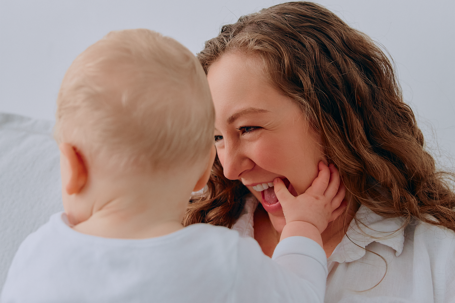 A smiling mother gazes lovingly at her baby, sharing a joyful moment together against a soft white background.