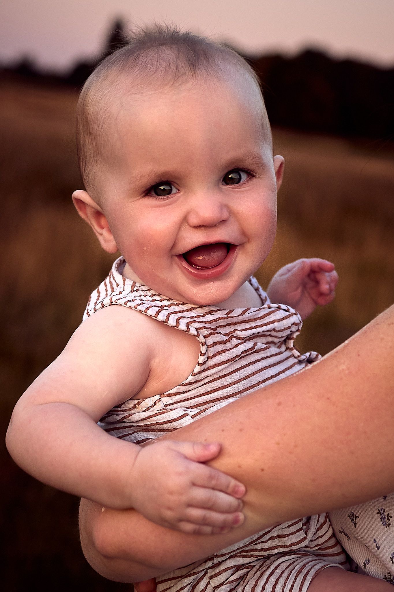 A happy baby in striped clothing smiles brightly during sunset.