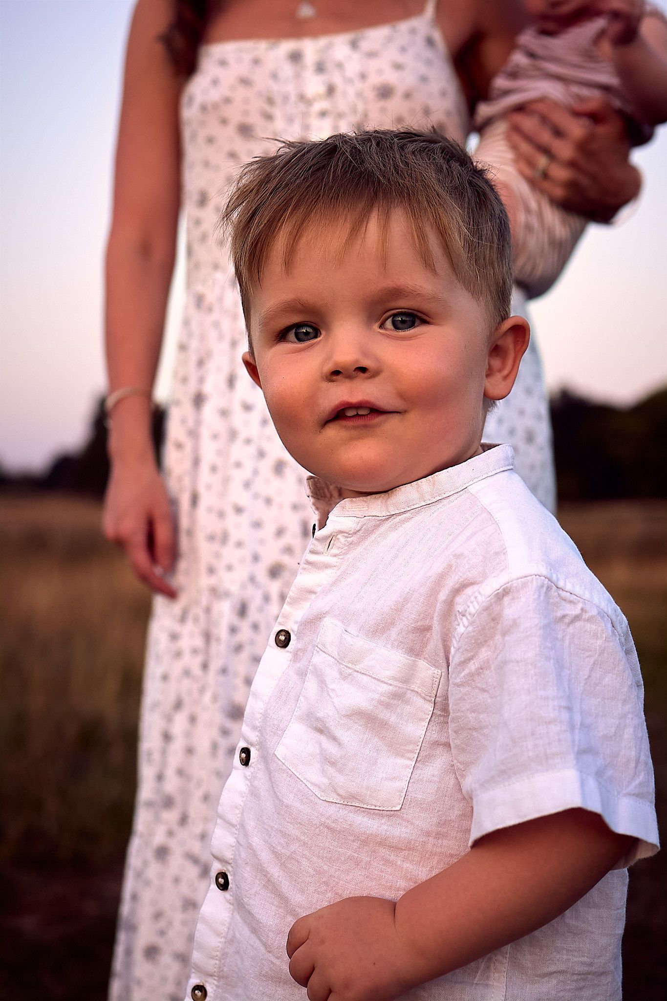 A young child in a white button-down shirt smiles at the camera during golden hour.