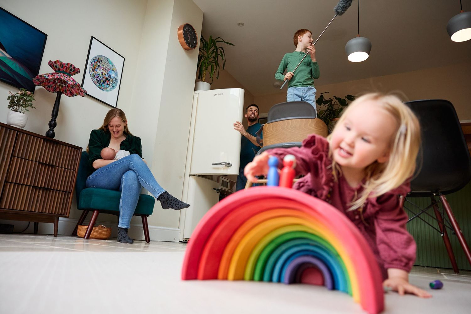 Family relaxing at home with toddler playing with colorful wooden rainbow toy on living room floor.