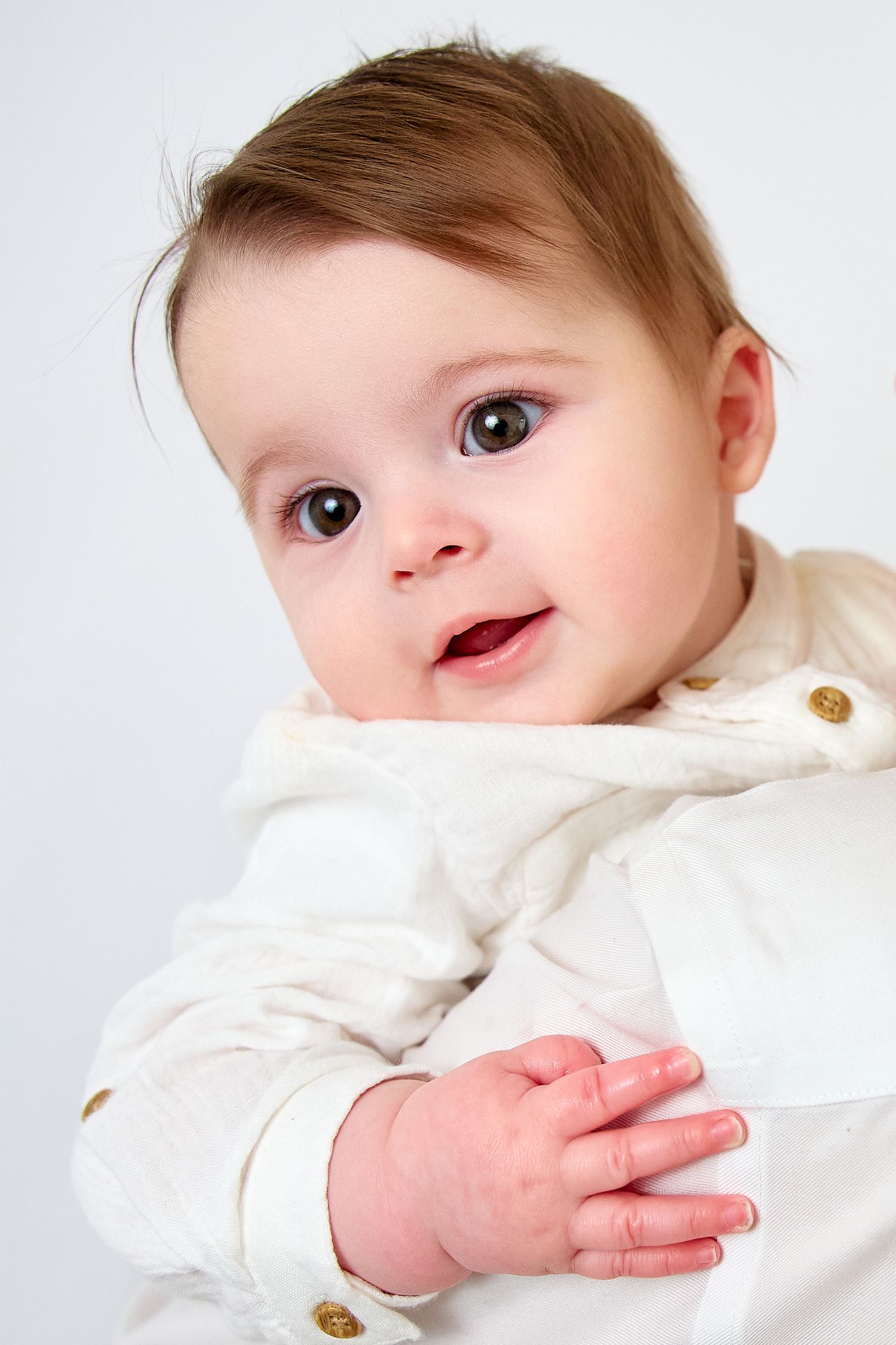 Adorable baby with big brown eyes wearing a white outfit, smiling and clasping tiny hands together.