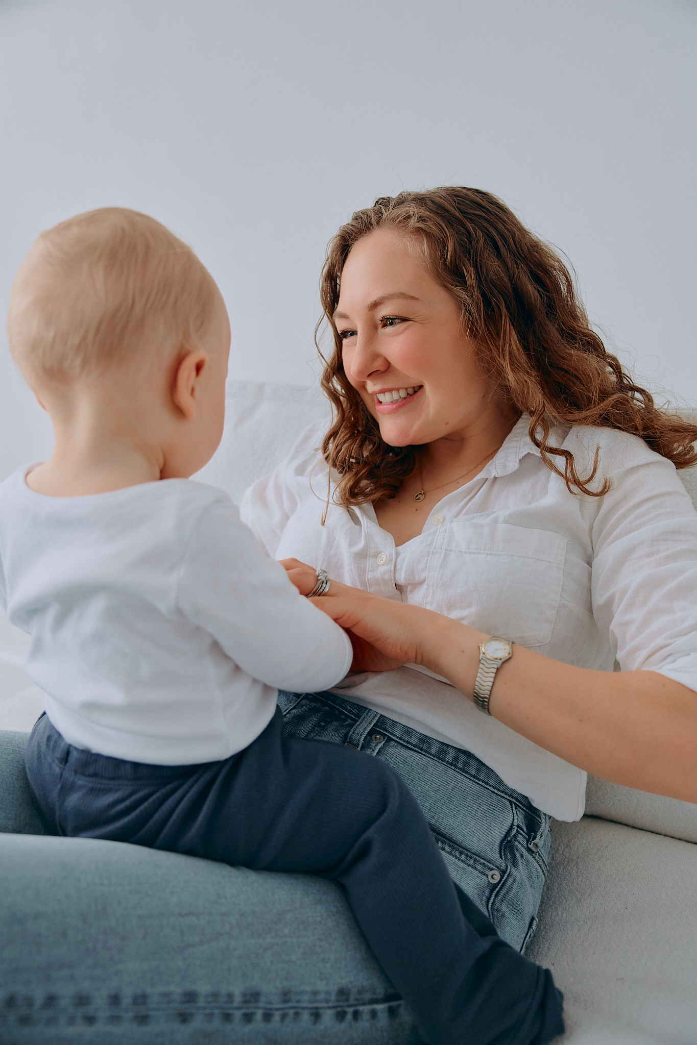Smiling mother in white shirt and jeans interacting with her baby on a white couch.