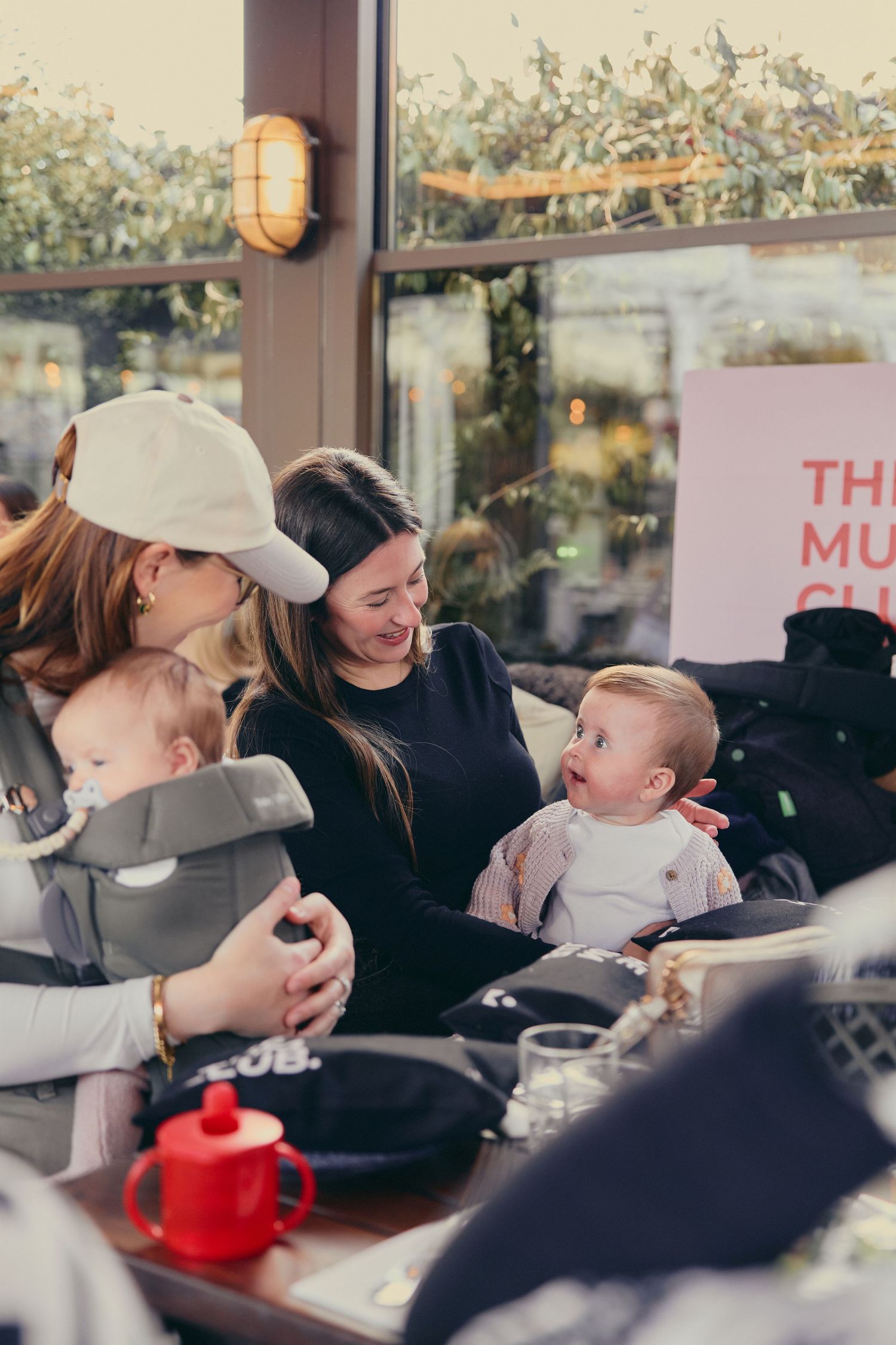 Two mothers smile at each other while holding their babies at a cozy cafe gathering.