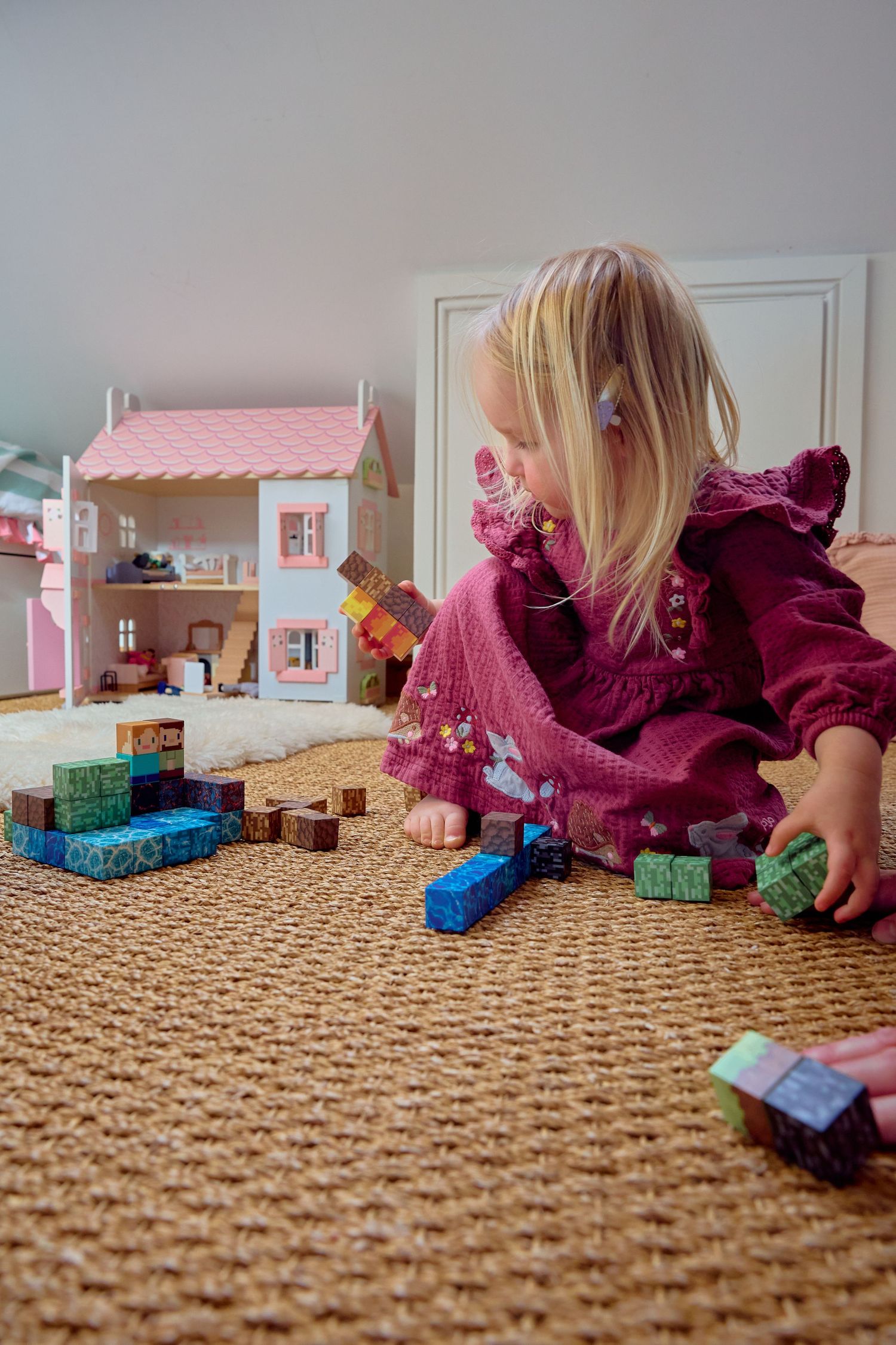 Young blonde girl in pink dress playing with wooden blocks and dollhouse on woven rug.