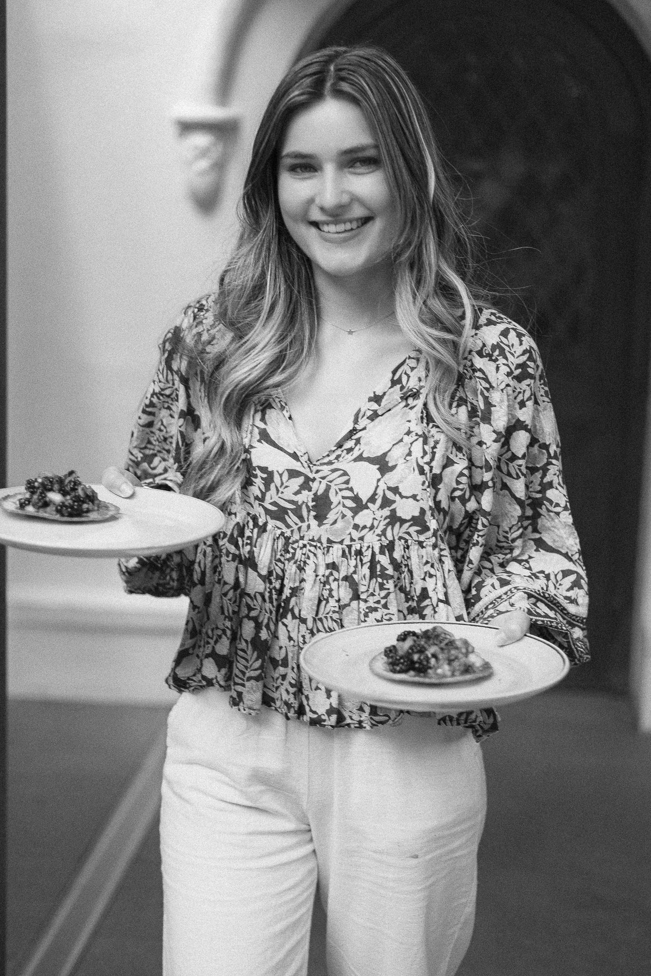 A person in a patterned top and white pants holds plates of food in a casual dining setting.