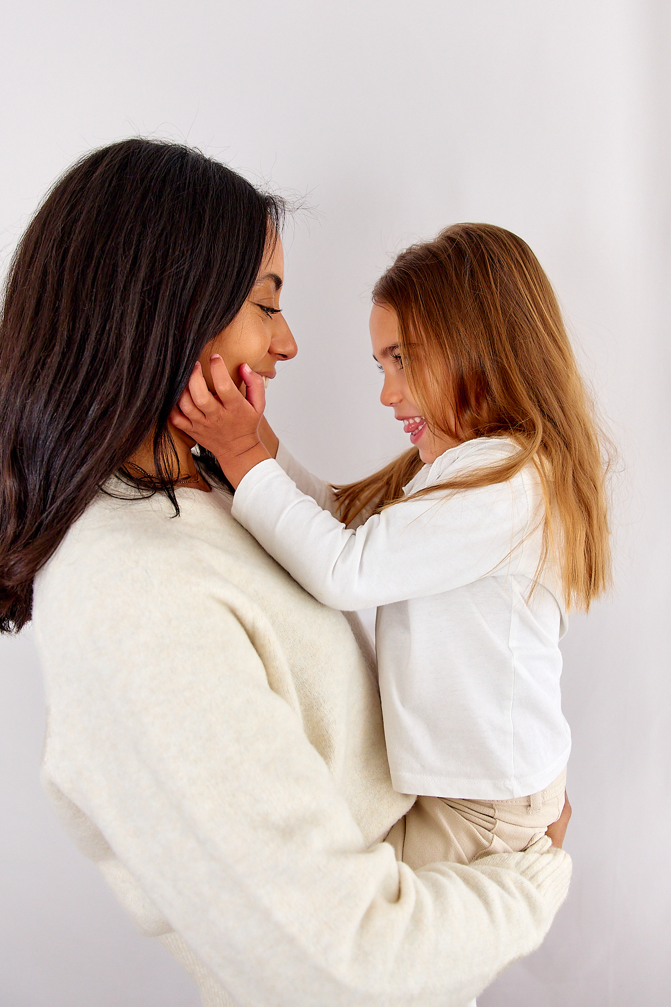 A smiling mother holds her young daughter in her arms, sharing a tender face-to-face moment in matching cream sweaters.