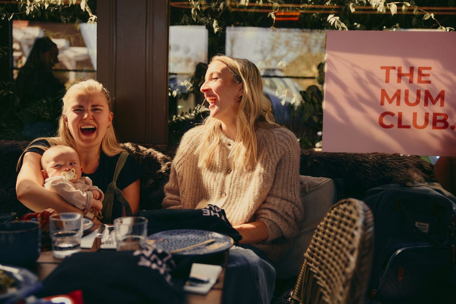 Two women laughing joyfully at The Mum Club event, one holding a baby.