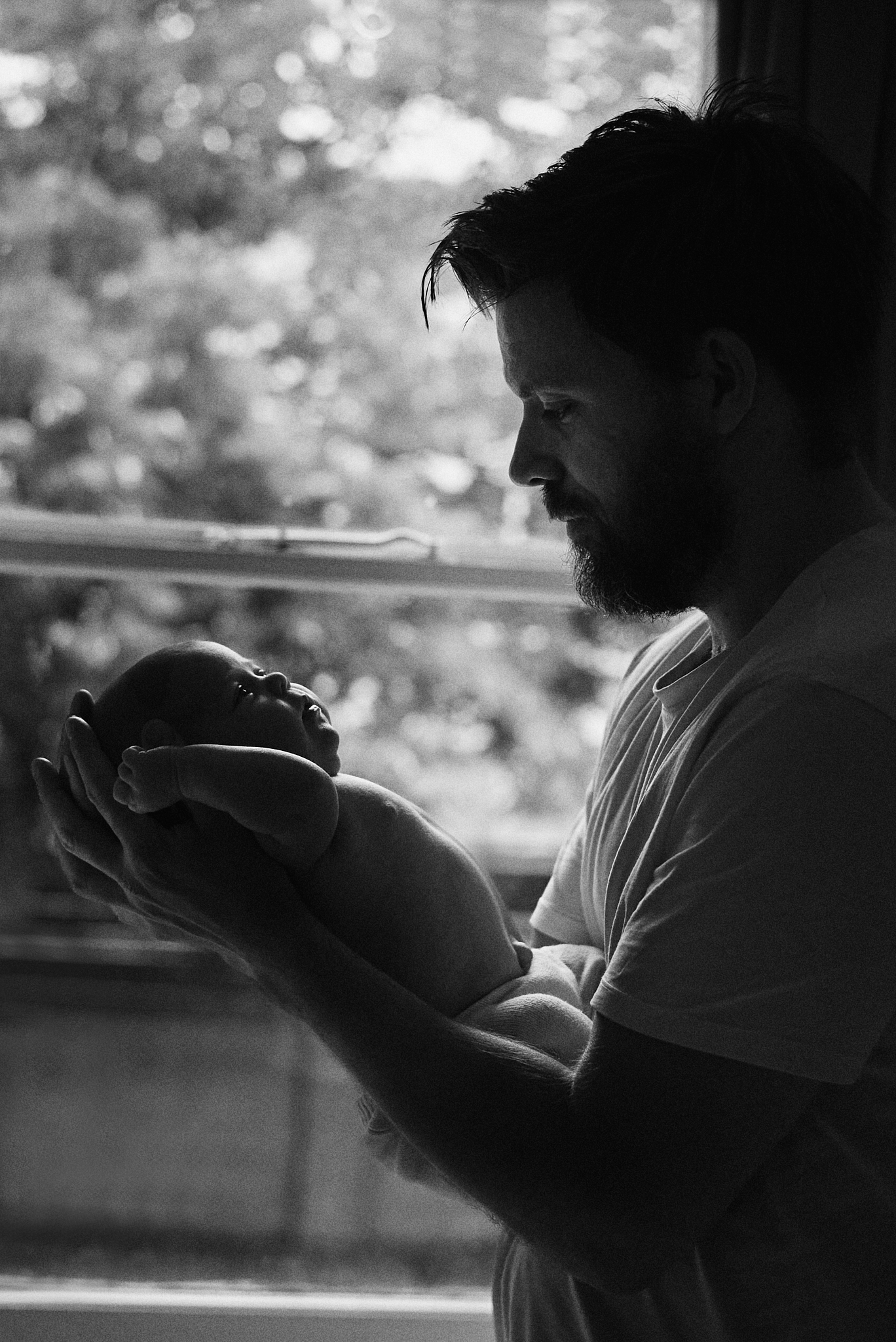 Black and white photo of a bearded father tenderly holding his newborn baby near a window.