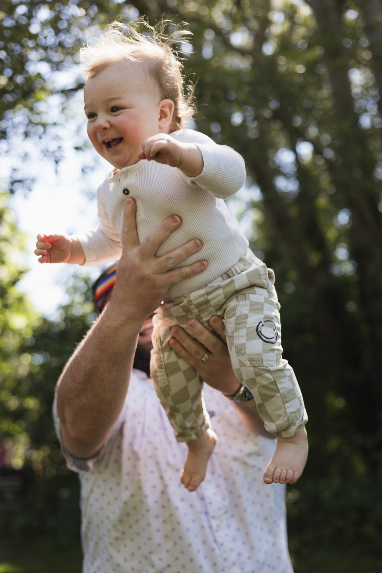 Baby in white outfit being playfully lifted and held in outdoor setting.